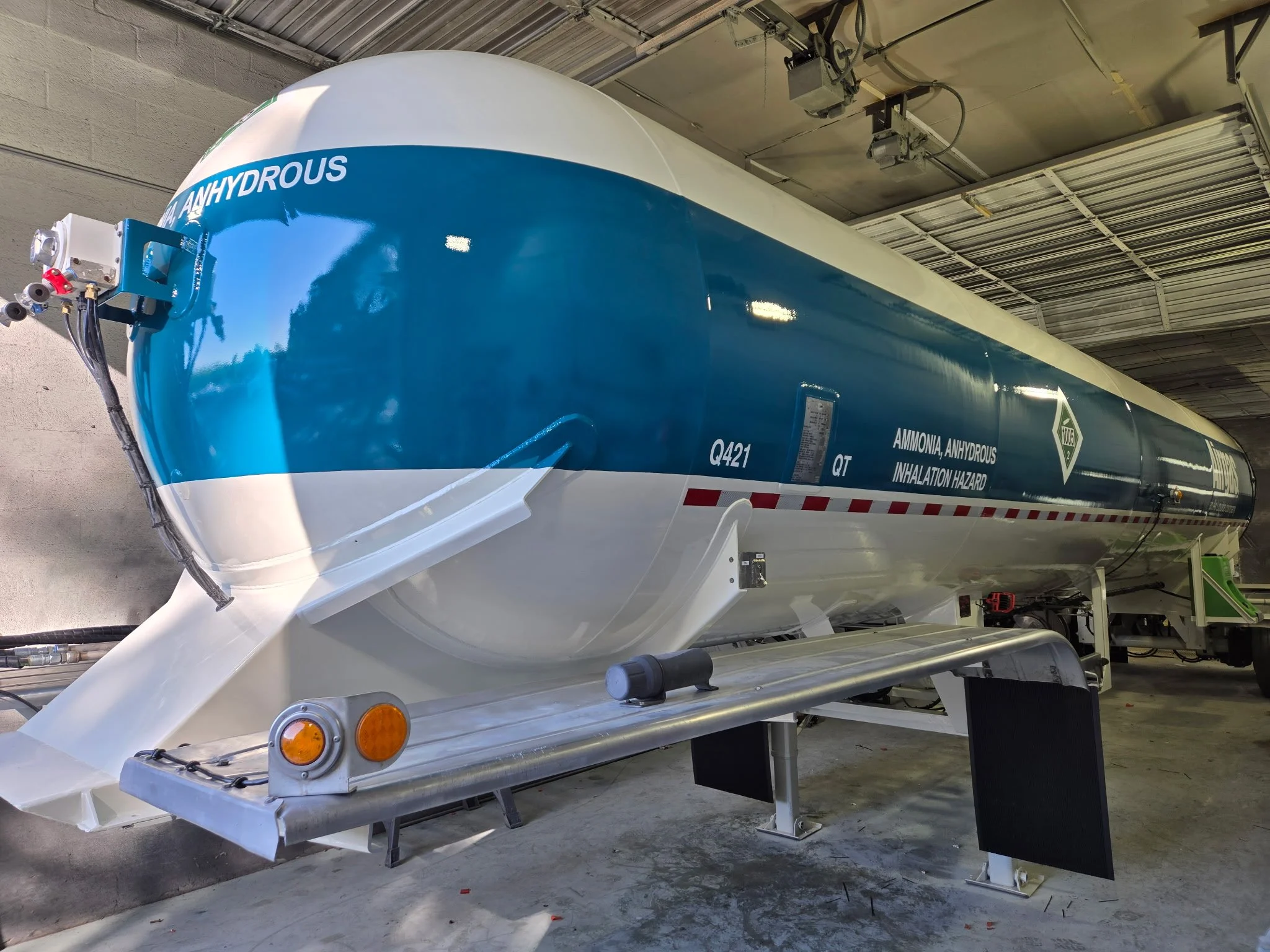 A large industrial tank labeled as containing ammonia and anhydrous, used for inhalation hazard, with indication of Q421 and QT. It is stored indoors, mounted on a support with warning lights and connected to hoses. It was used for a body paint job.