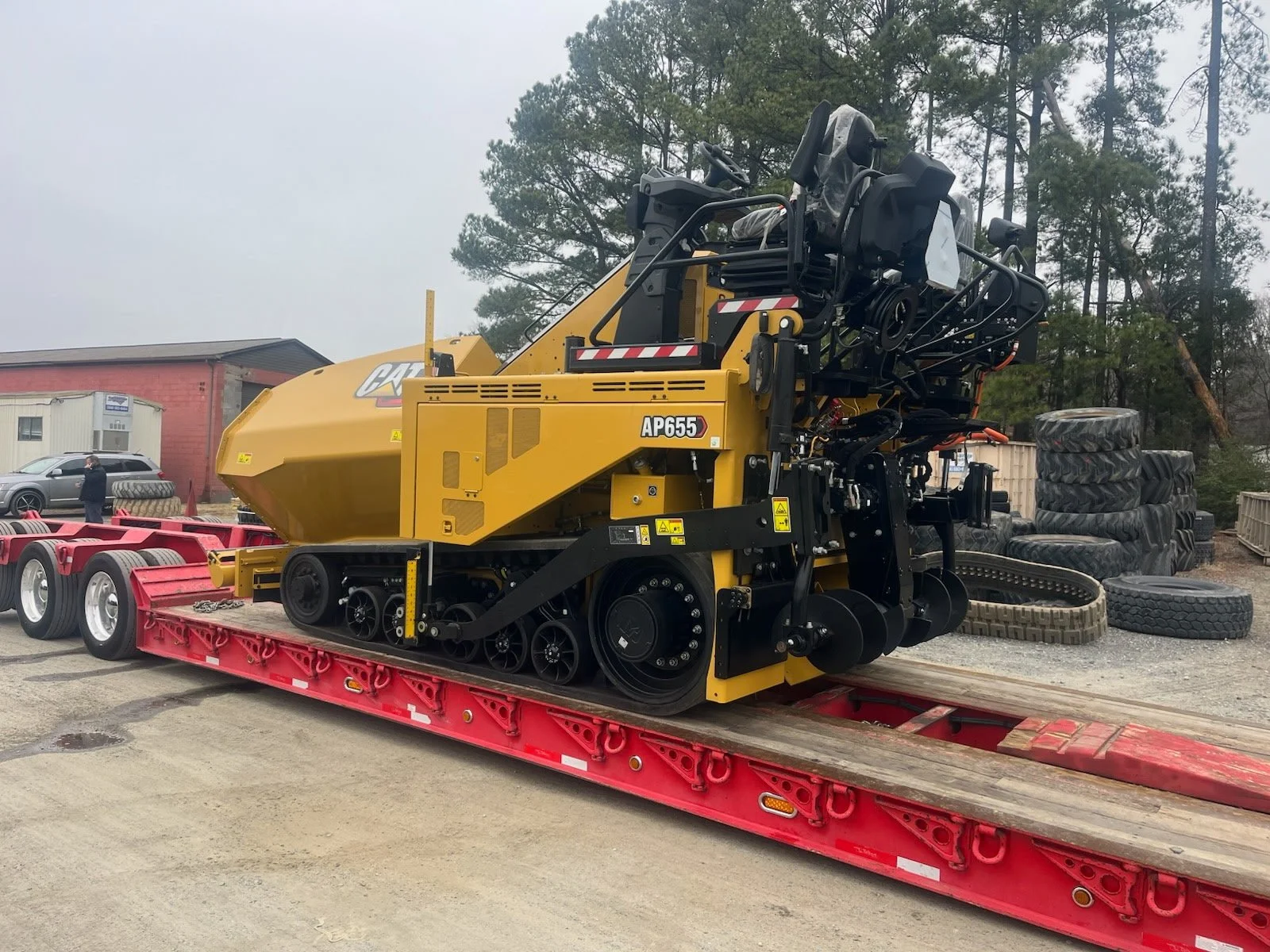 Large yellow CAT AP655 dozer on a red flatbed trailer, with a gray sky and stacks of tires in the background. Body paint job for heavy equipment.