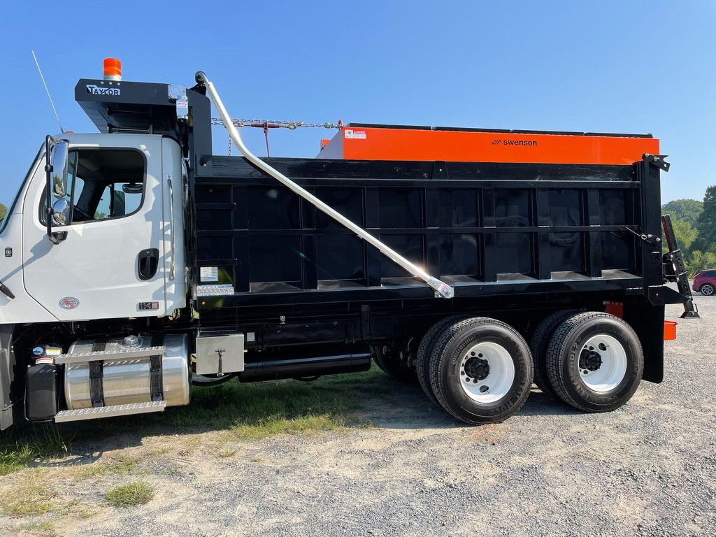 Side view of a dump truck with a black and orange bed parked on a gravel lot under a clear blue sky. Paint job for a used heavy truck company.
