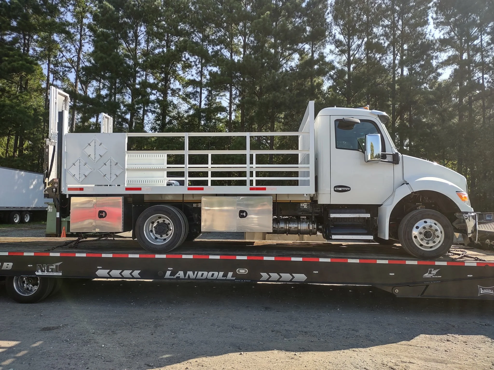 White flatbed truck on a trailer with trees in the background. Body paint and body repair for a used heavy truck company.