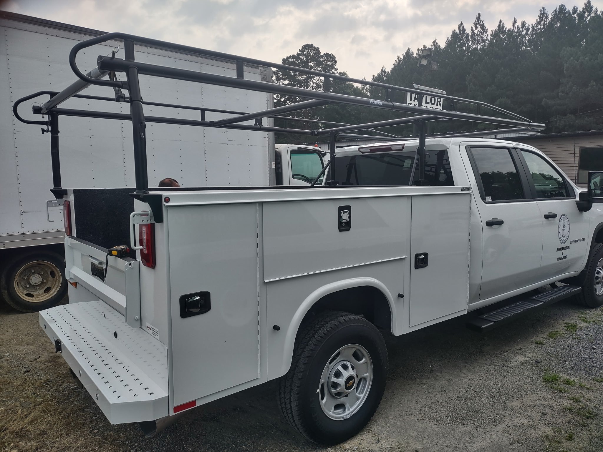 White utility truck with a ladder rack on top, parked outdoors next to a white box truck and a building in the background. Body paint and body repair for a used heavy truck fleet company.