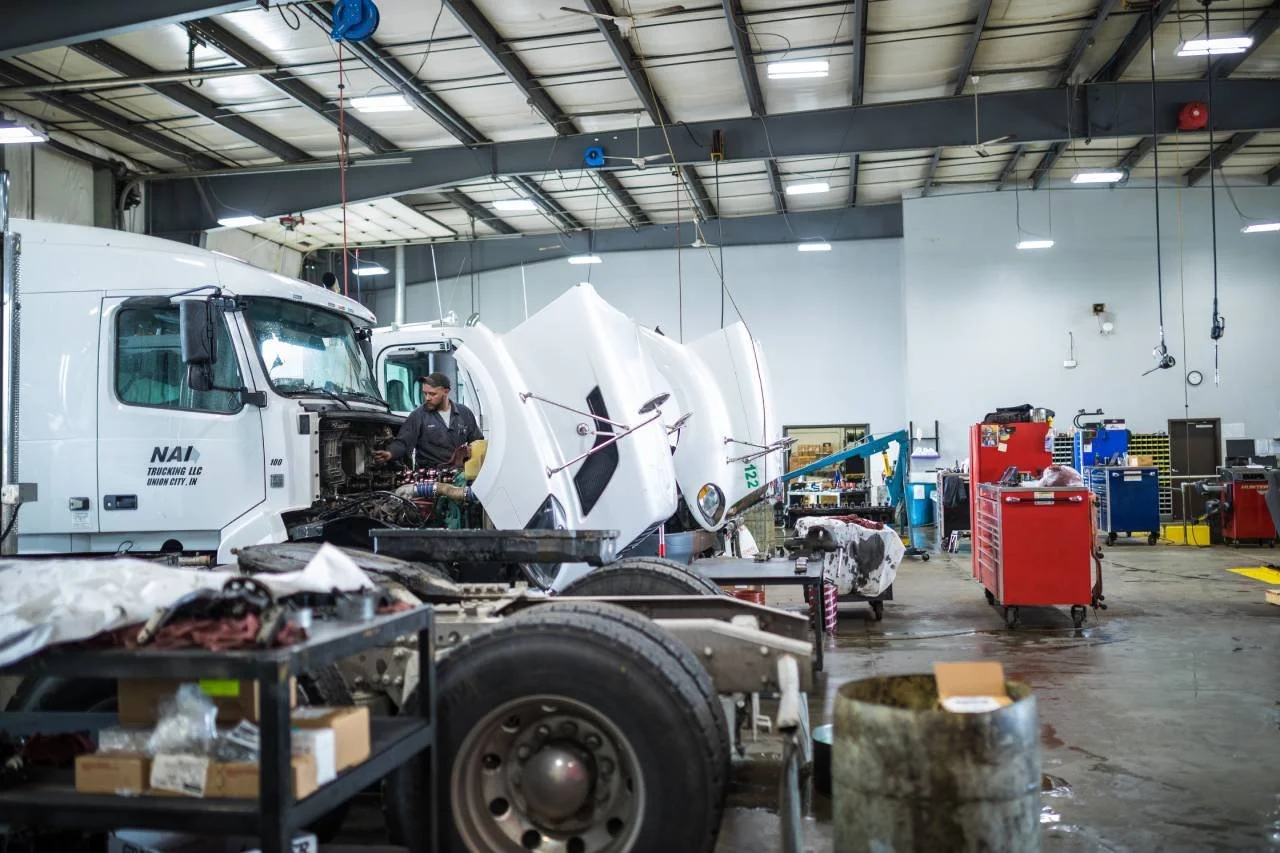 Inside an auto repair shop with a white truck undergoing repairs. The truck's front panel is removed, and a mechanic is working on the engine. The shop has tools and equipment, including red tool carts and workbenches, with a high ceiling and overhead lights.