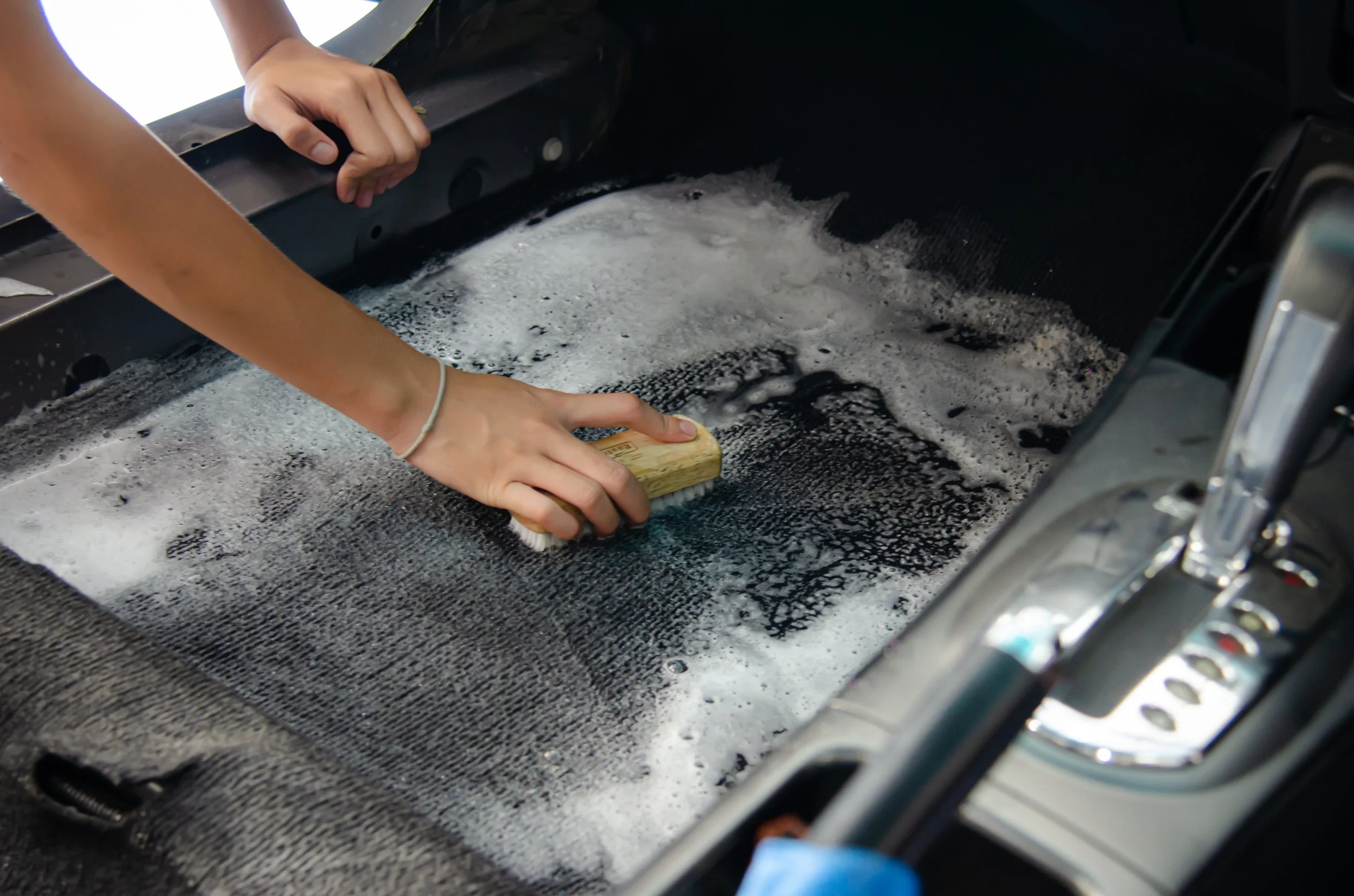 Person cleaning the interior floor of a car with a brush and soap and water.