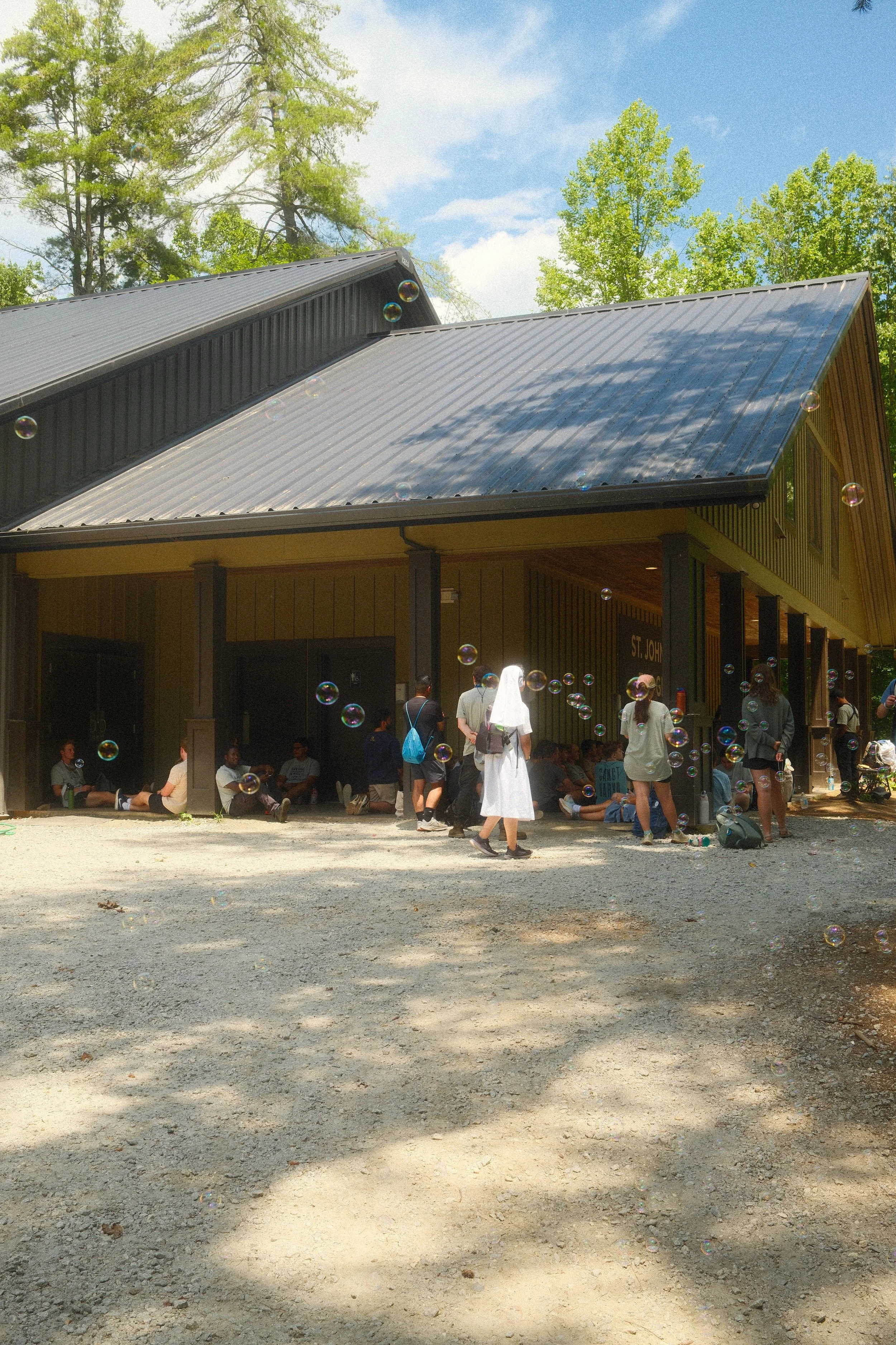 People waiting outside a wooden building on a sunny day with some sitting and some standing, surrounded by trees and soap bubbles.