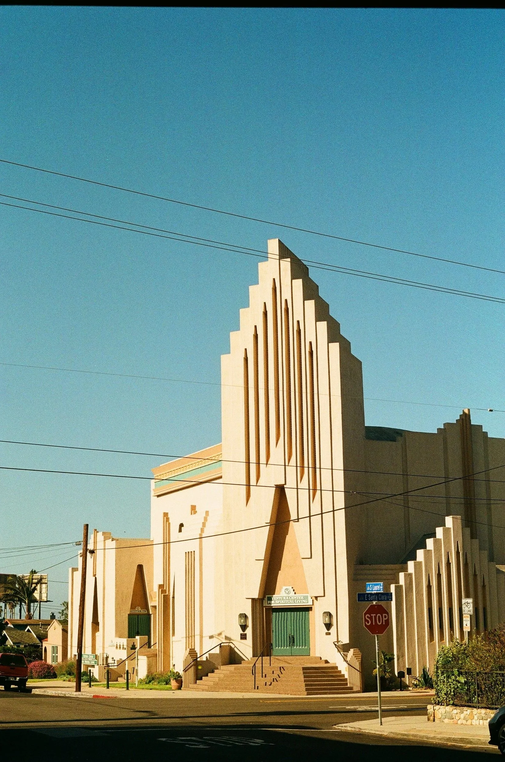 A modern church with art deco architectural style, featuring tall, narrow windows and geometric design, located at a street corner with a stop sign, power lines, and clear blue sky in the background.