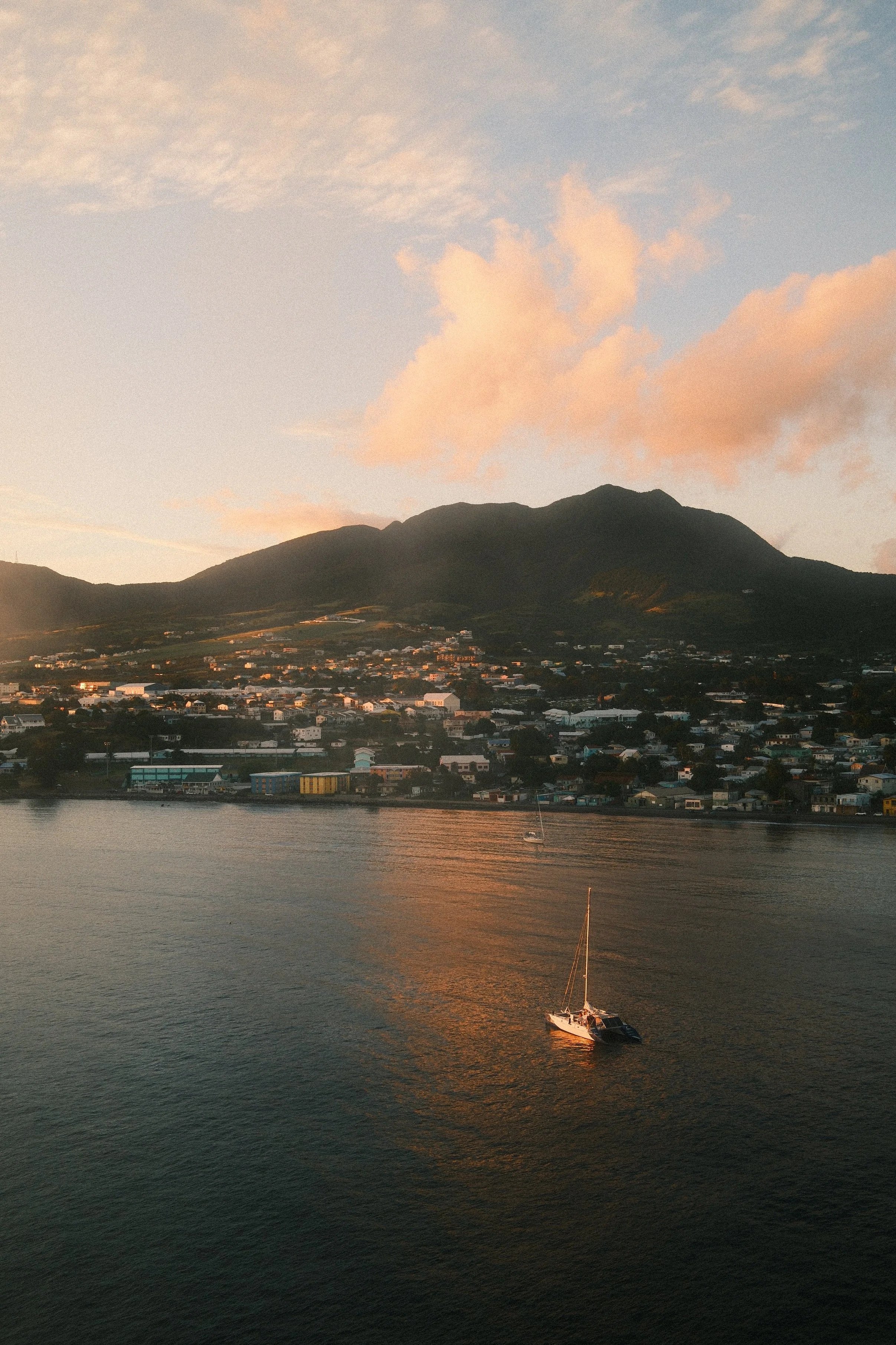 A sailboat floating on calm water with a lakeside town and a mountain in the background during sunset.