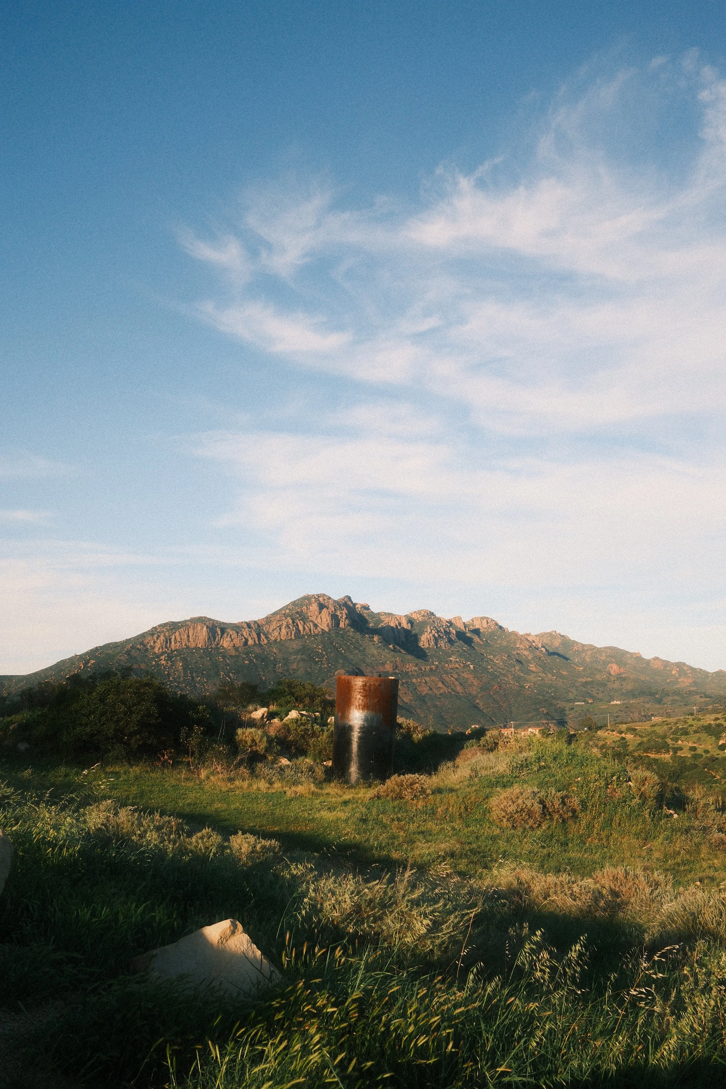 Landscape with green grass, a mountain range in the background, and a blue sky with wispy clouds. A rusty metal barrel is in the foreground.