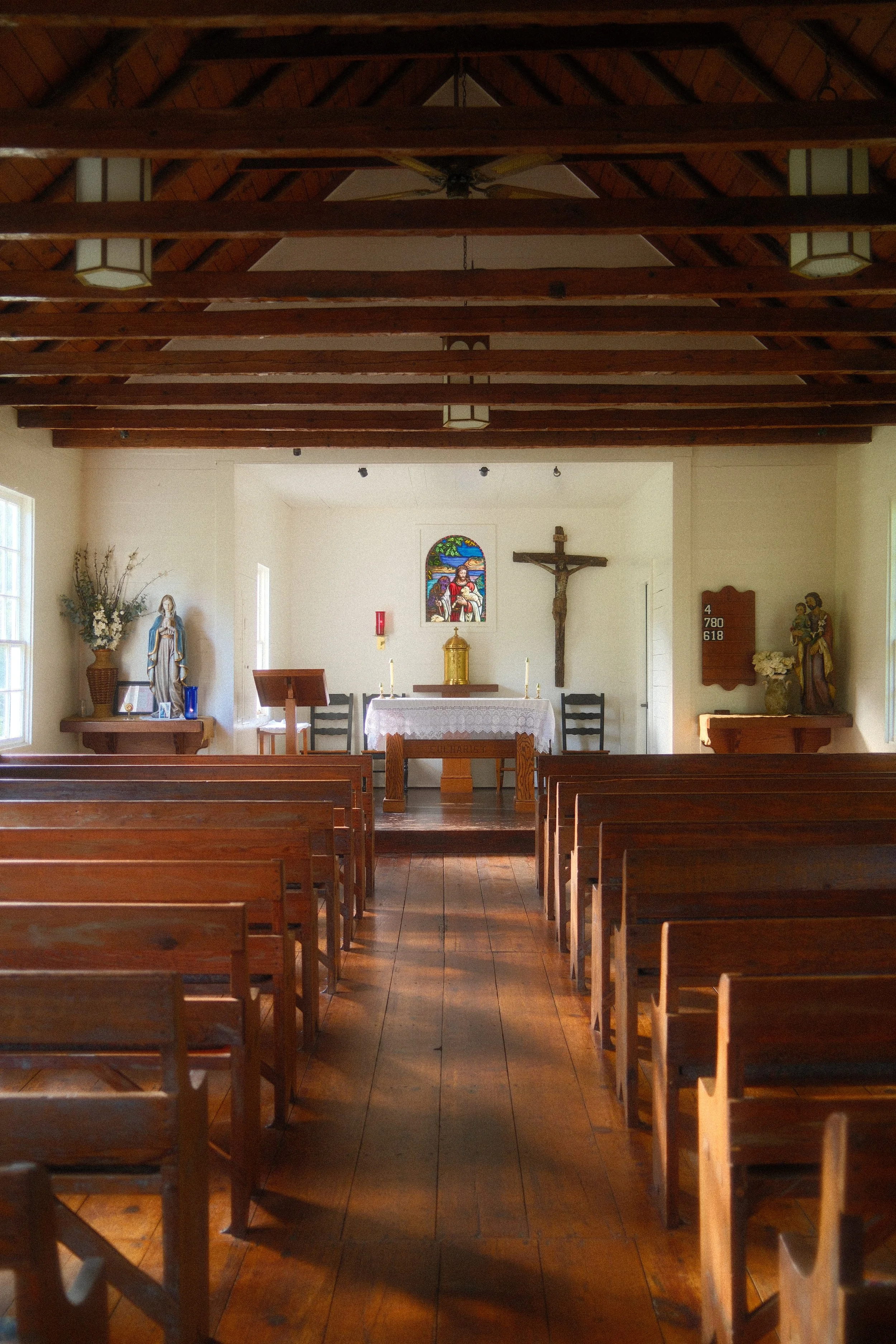 Interior of a small church with wooden pews facing an altar with a crucifix, statue, and stained glass window.