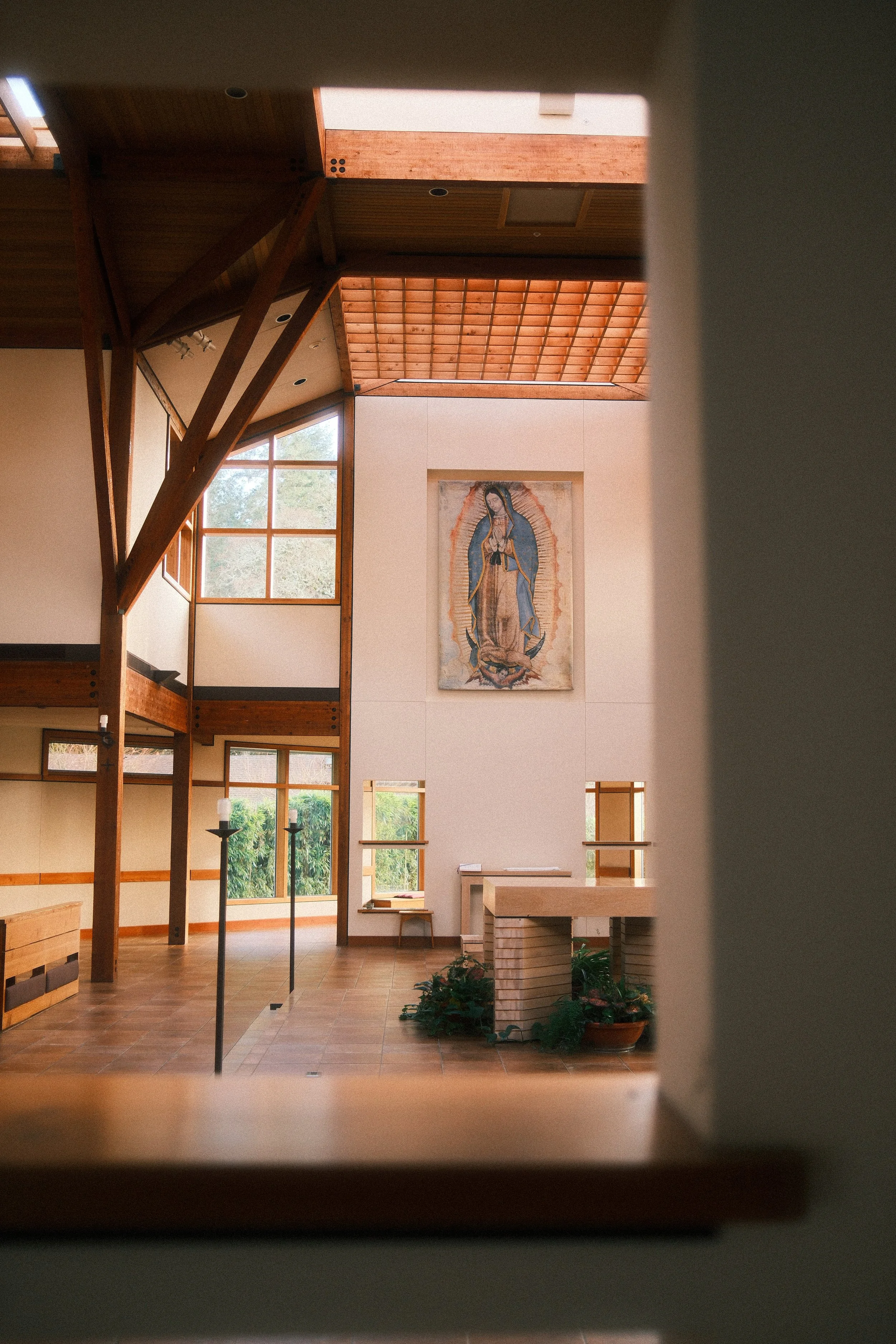 Interior of a church with wooden beams, large windows, and a painting of the Virgin of Guadalupe on the wall.