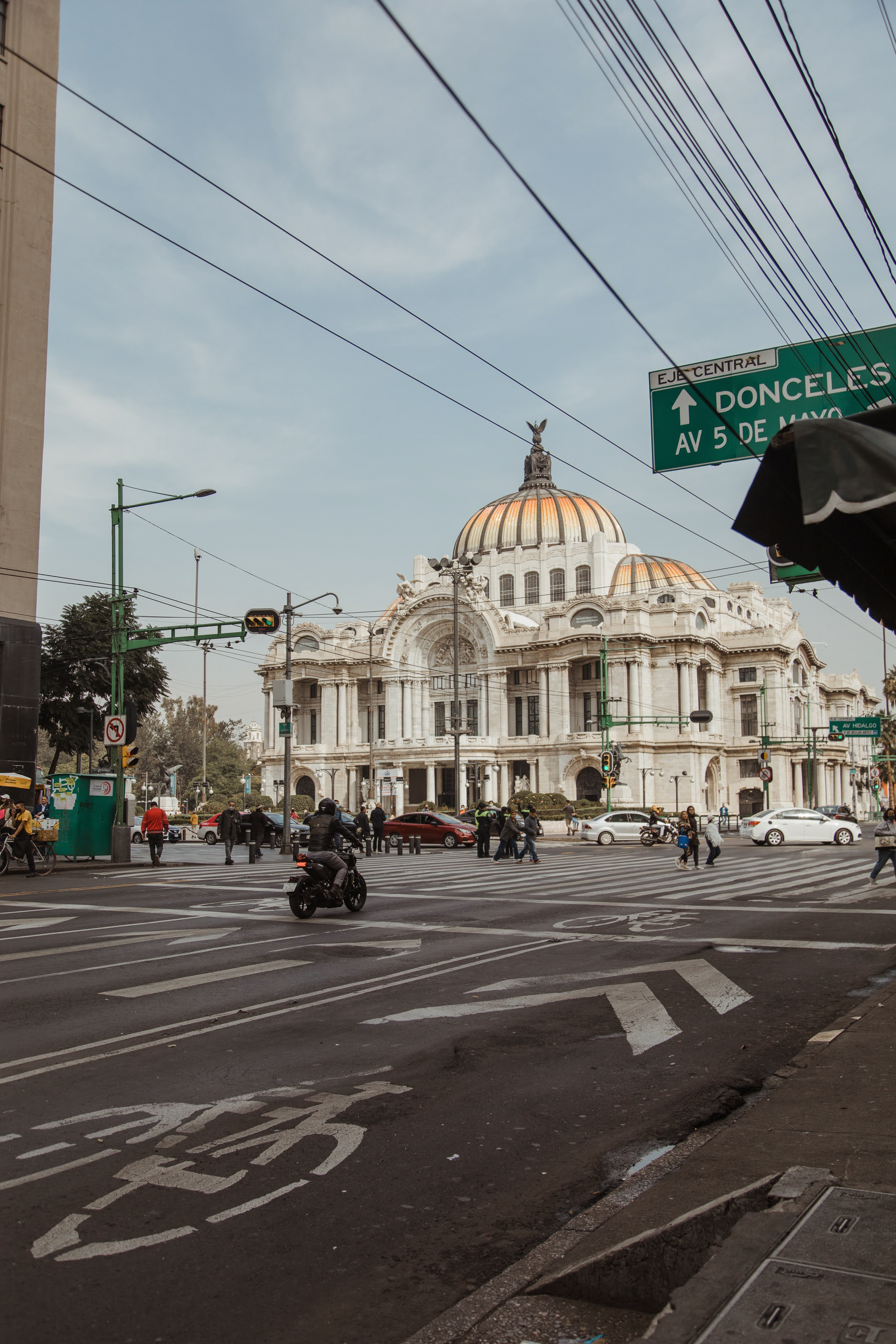 Street scene in Mexico City with the Palacio de Bellas Artes in the background, busy crosswalk with pedestrians, cars, and a motorcyclist, overhead street signs, power lines, and traffic signals.