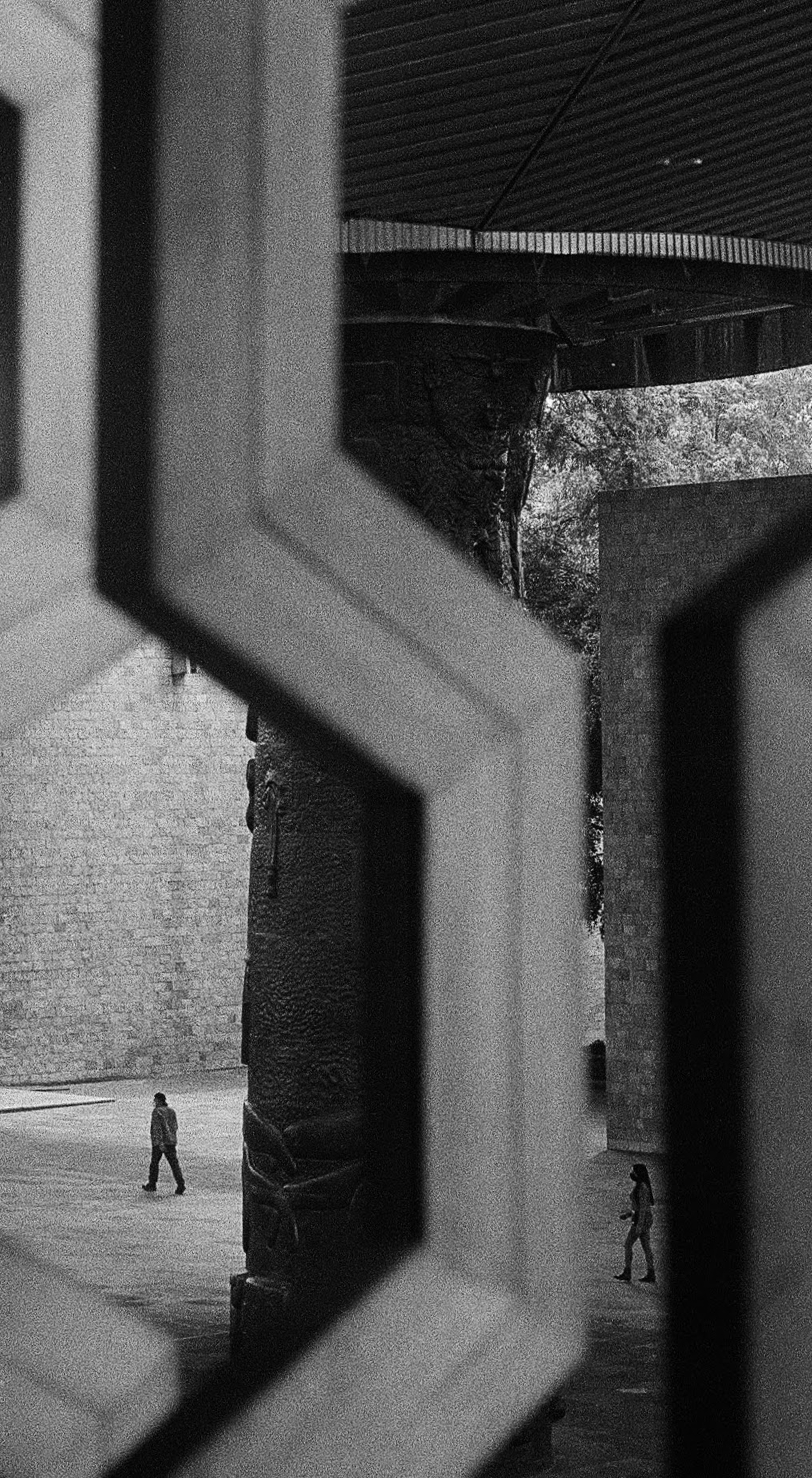 Black and white photo showing two people walking in opposite directions, seen through a geometric pattern of a fence or railing. One person is on the left side, and the other on the right side of the image, with an urban background including a wall a