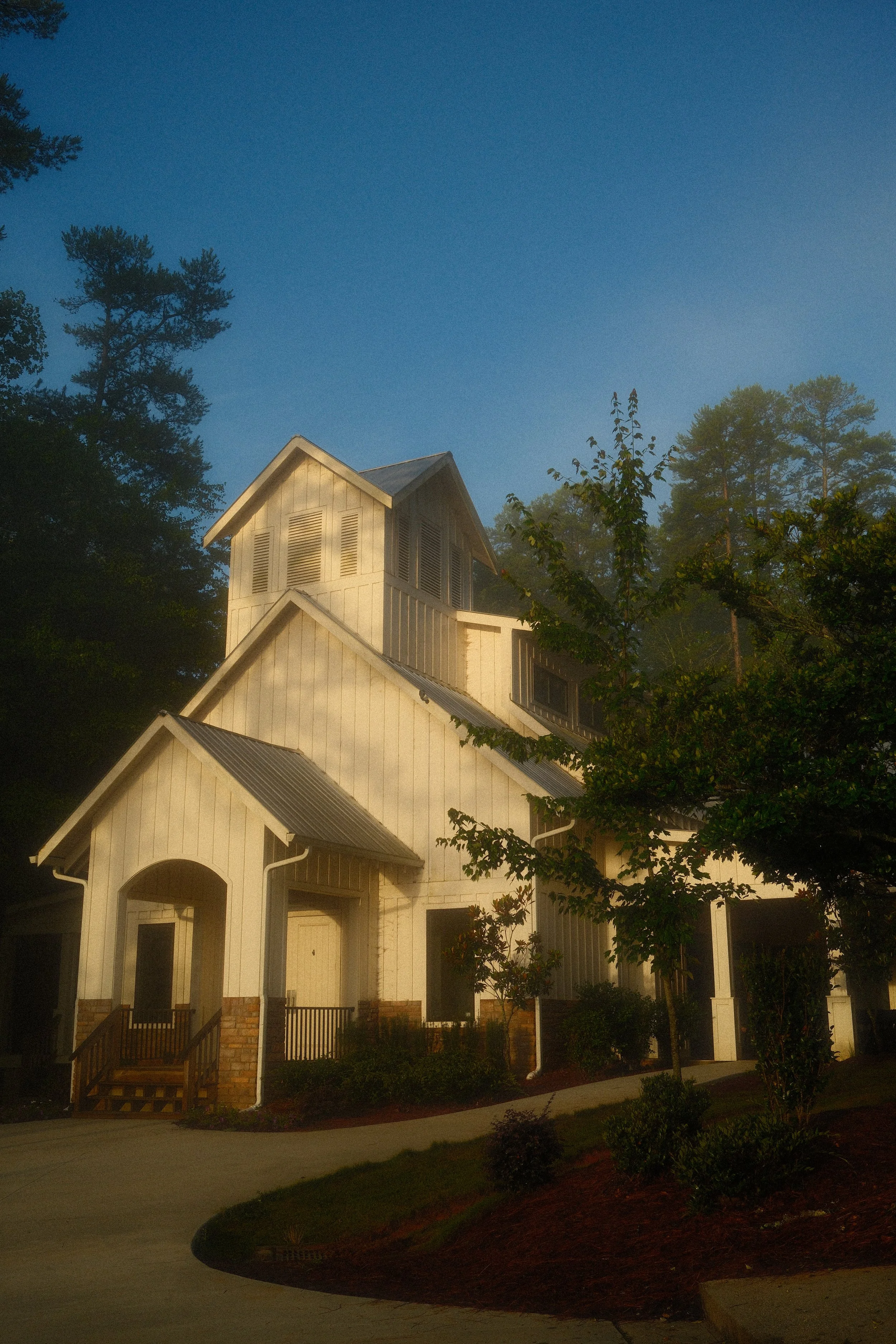 A white, multi-story house with a steep roof, surrounded by trees and a landscaped yard, under a clear blue sky.