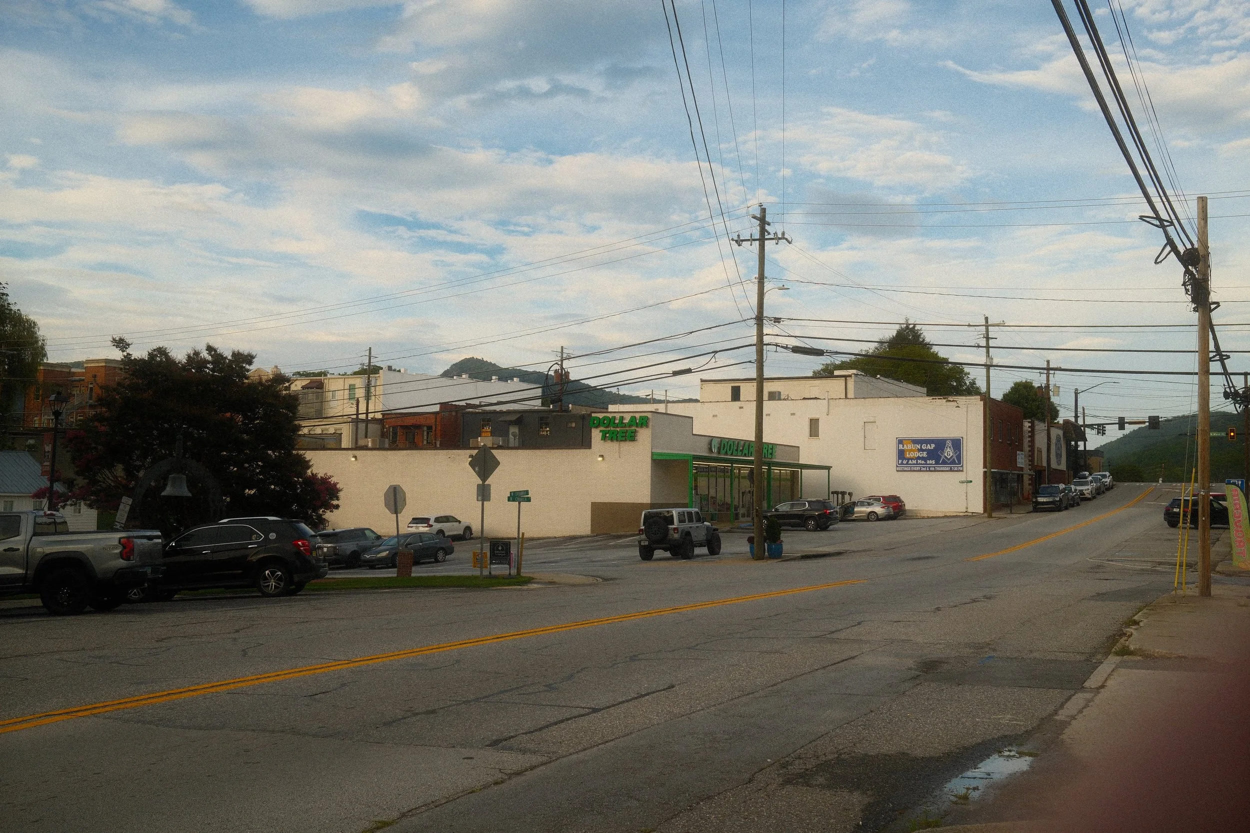 A street view showing parked cars in front of a convenience store named Dollar Tree, with residential and commercial buildings in the background, utility poles, and a cloudy sky.