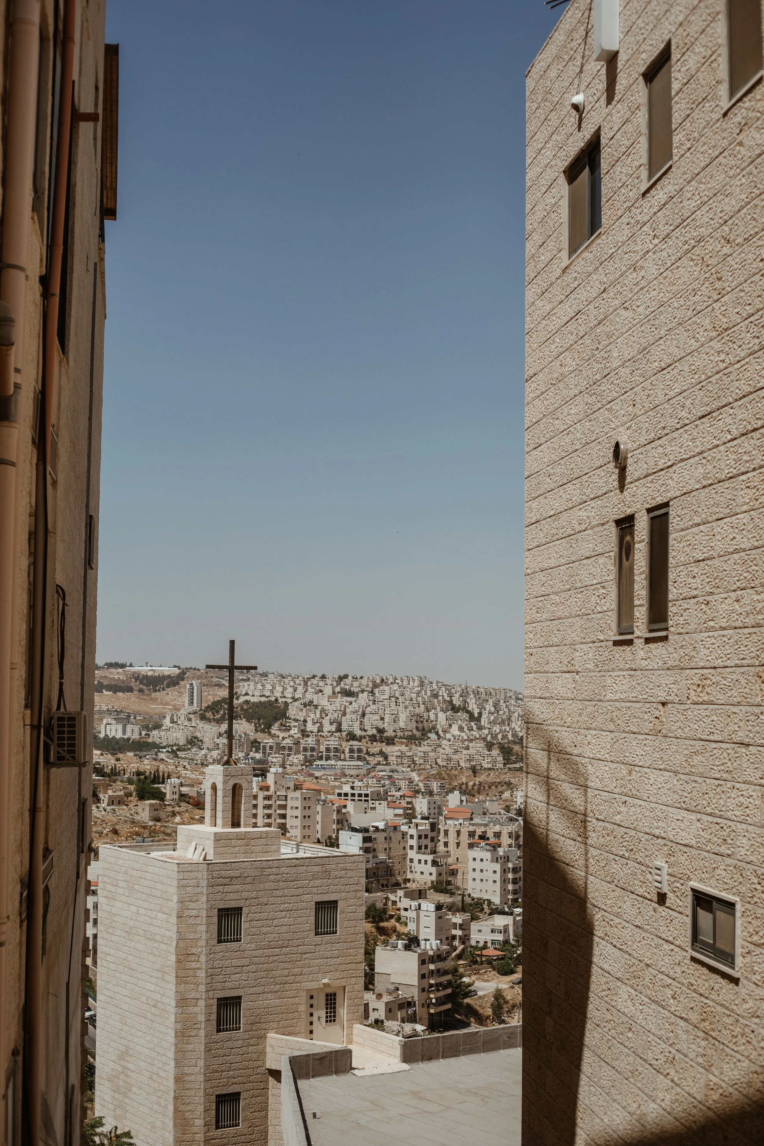 City view with a church and a cross on a building, seen through a narrow gap between two buildings with many residential buildings on a hillside in the distance, under a clear blue sky.