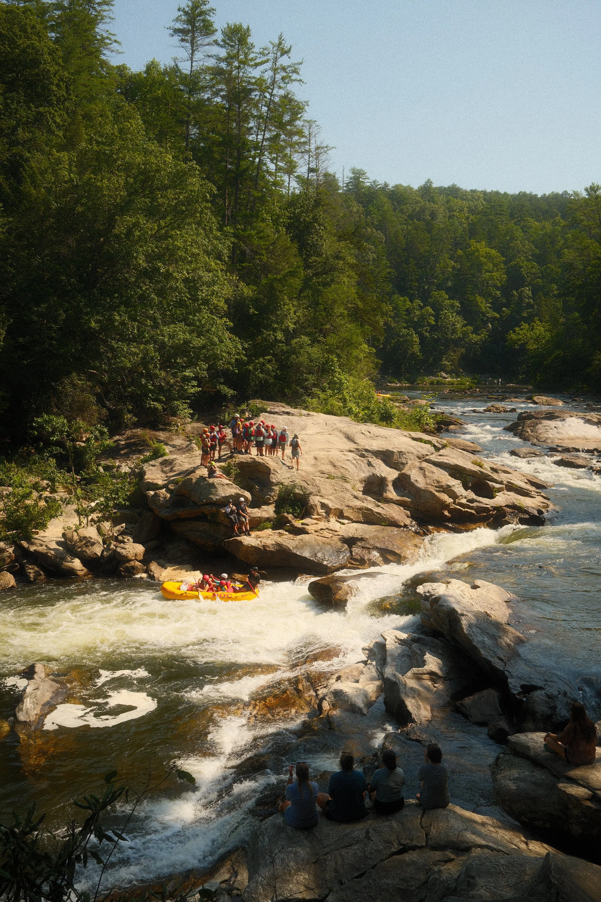 People rafting on a river surrounded by rocks and trees, with some spectators sitting on rocks nearby, under a clear sky.