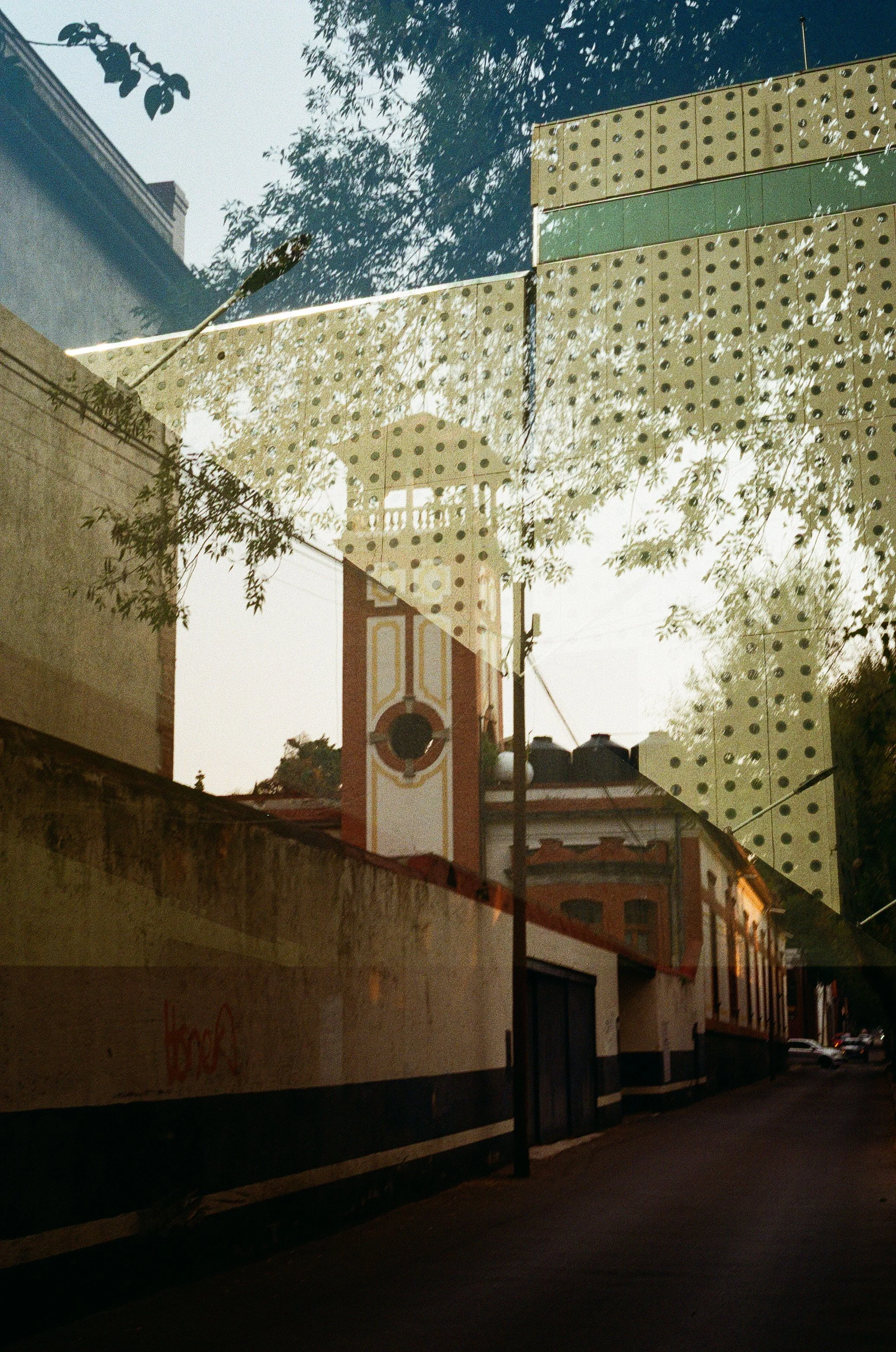 Reflected view of a historic building with a clock tower in a glass window, with trees and a street scene in the reflection.