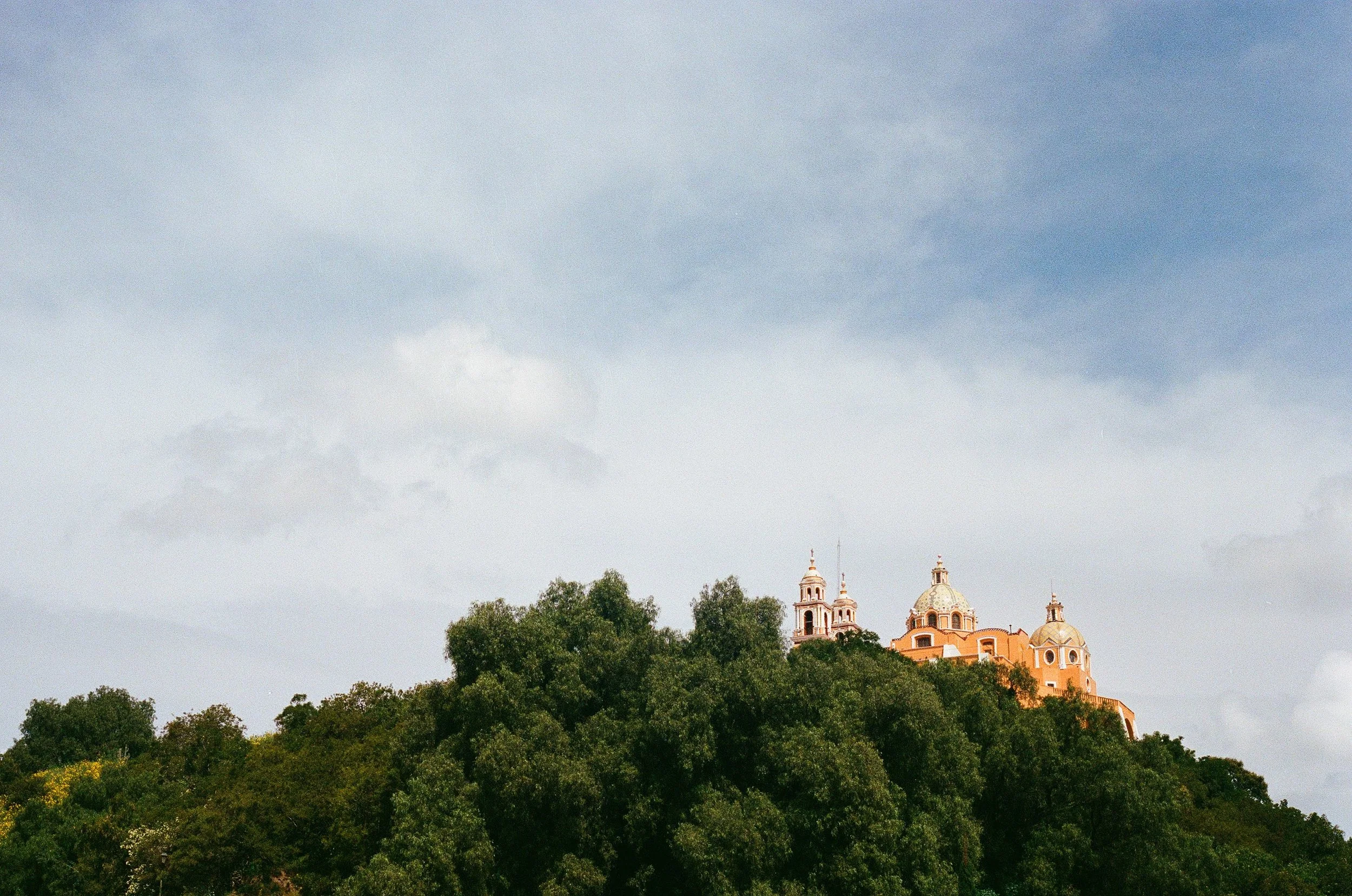 A historic building with multiple domes and towers on a hilltop surrounded by lush green trees against a cloudy sky.