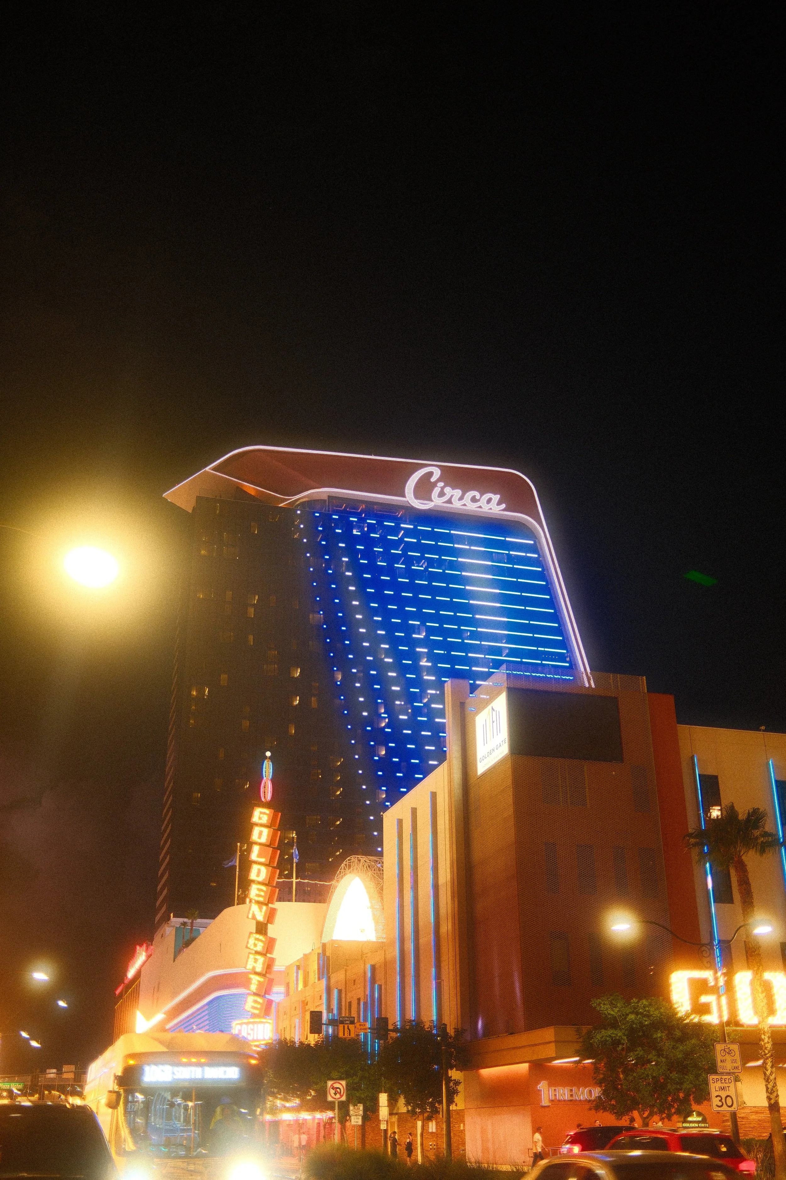 Nighttime scene of the Circa hotel with neon lights and signage, including the Golden Gate Casino sign, on a busy street with cars and pedestrians.