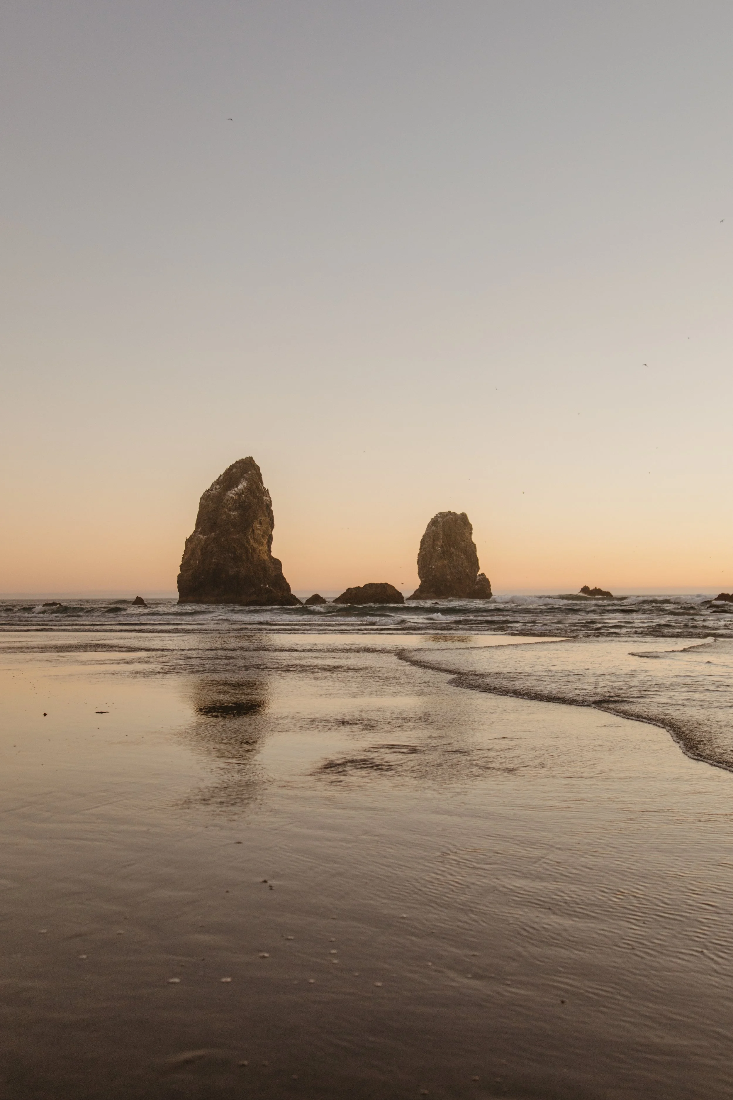 A beach at sunset with two large rock formations in the ocean and their reflections in the wet sand.