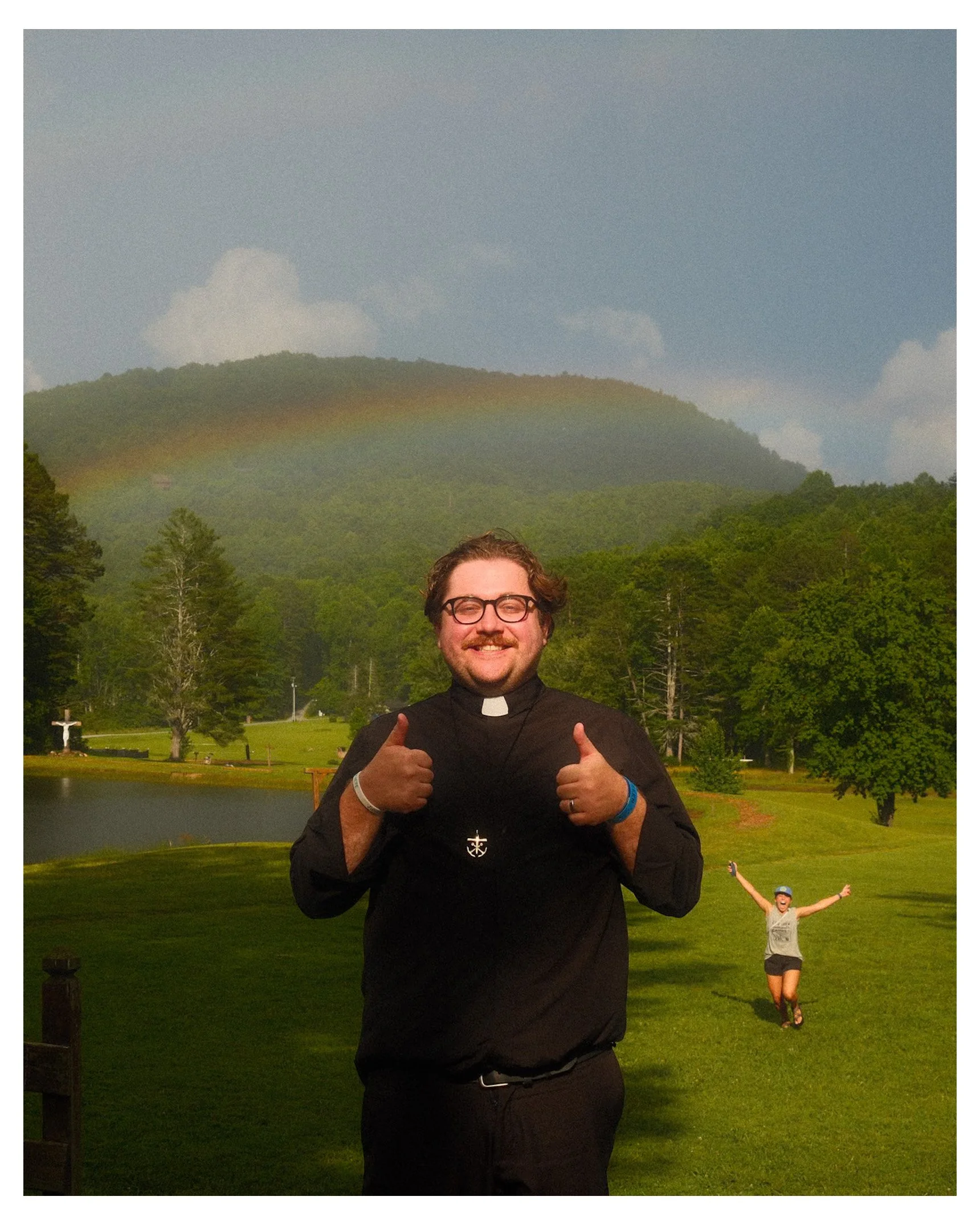 A smiling man dressed as a priest giving two thumbs up in a park with a lake, trees, mountains, and a rainbow in the background. A woman in the background is running with arms raised.