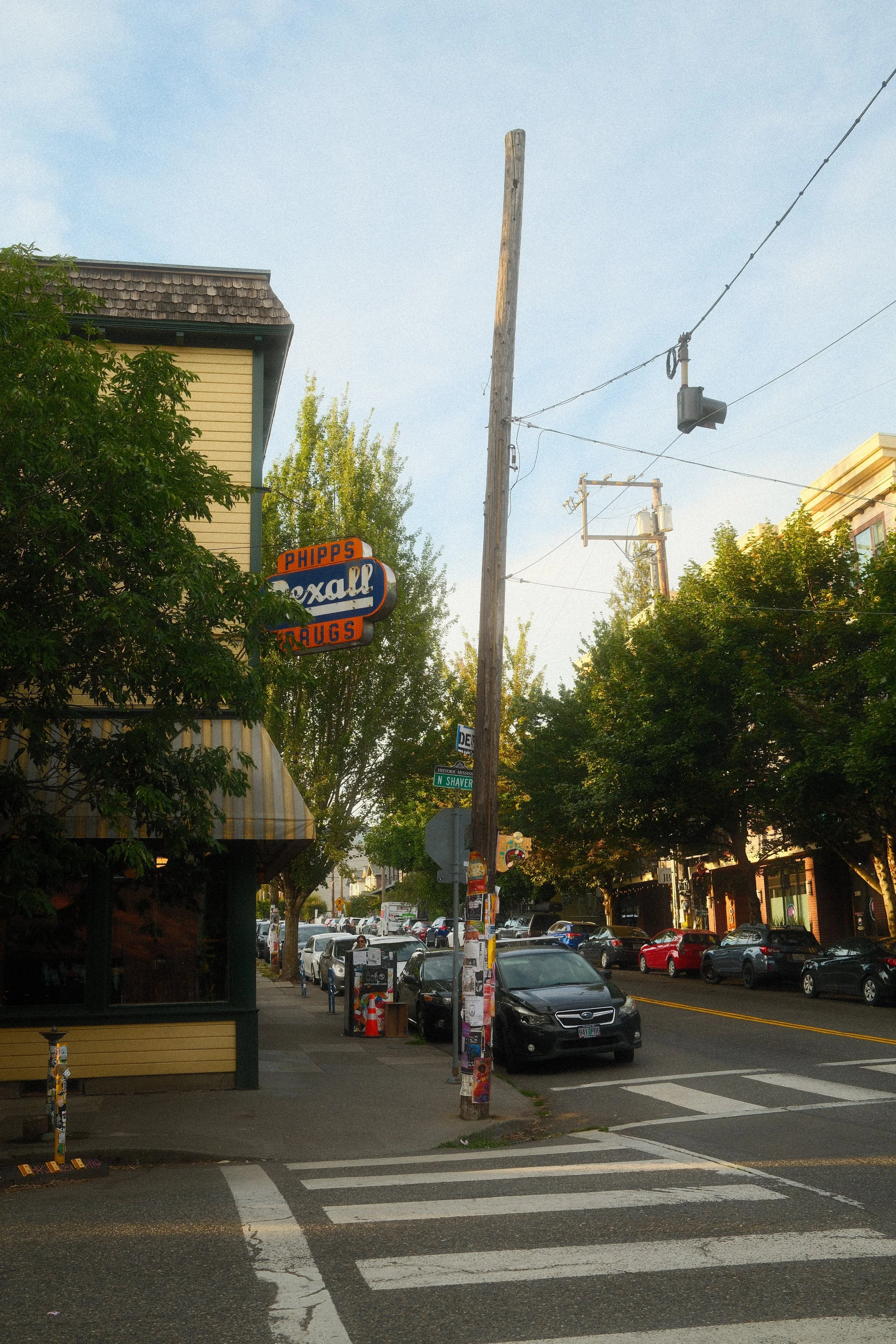 Street view with parked cars, a utility pole, and storefronts including a pharmacy called Phipps Rexall Drugs.