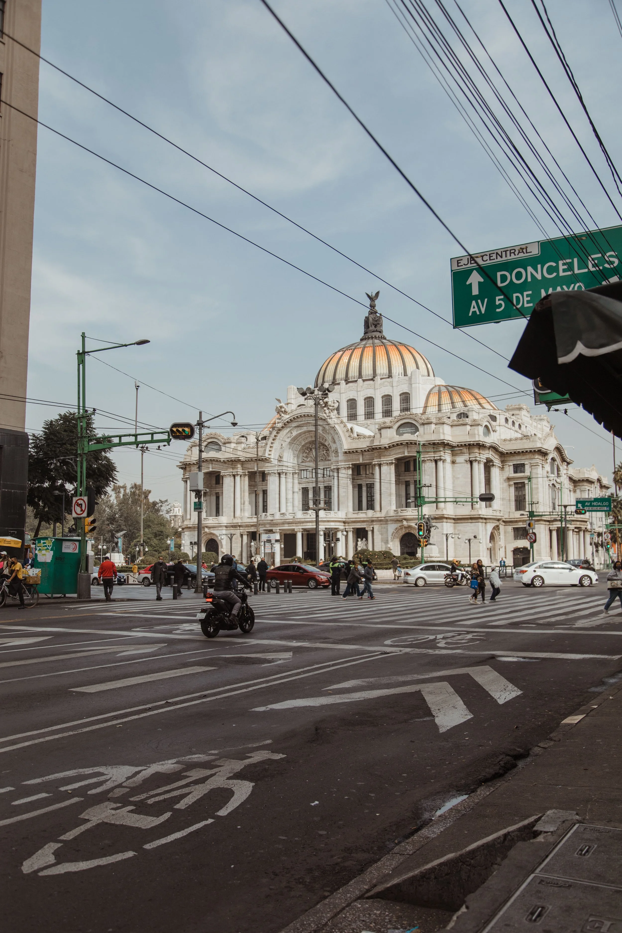 Street scene with a historic white building with a domed roof and sculptures, traffic lights, street signs, and people crossing the street in Mexico City.