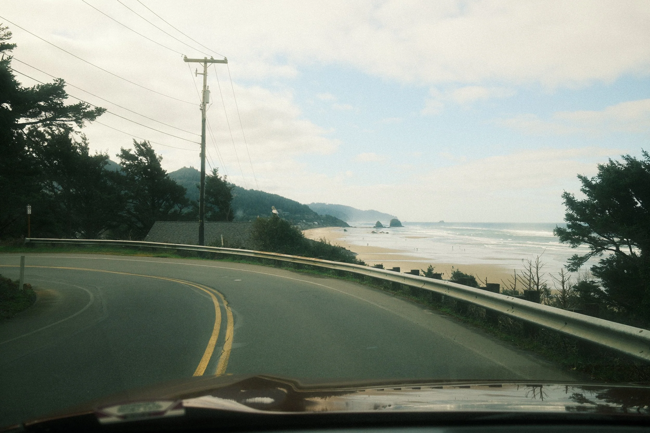A coastal road curves along a hillside with trees and power lines, overlooking a beach and ocean with rock formations in the distance on a cloudy day.