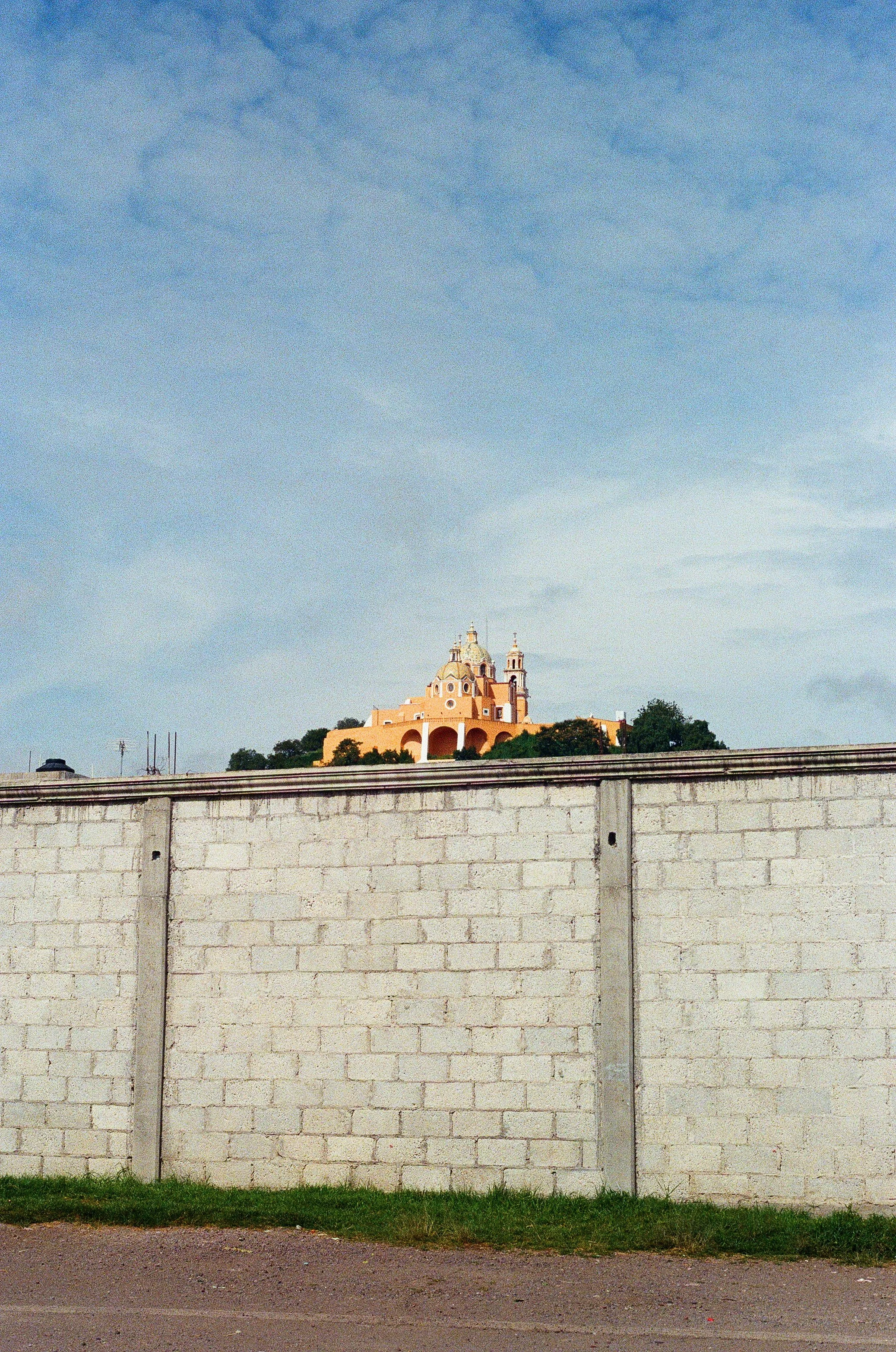 A church with beige and white architecture, situated on top of a hill, seen above a white brick wall with a grassy foreground and a blue sky with scattered clouds.