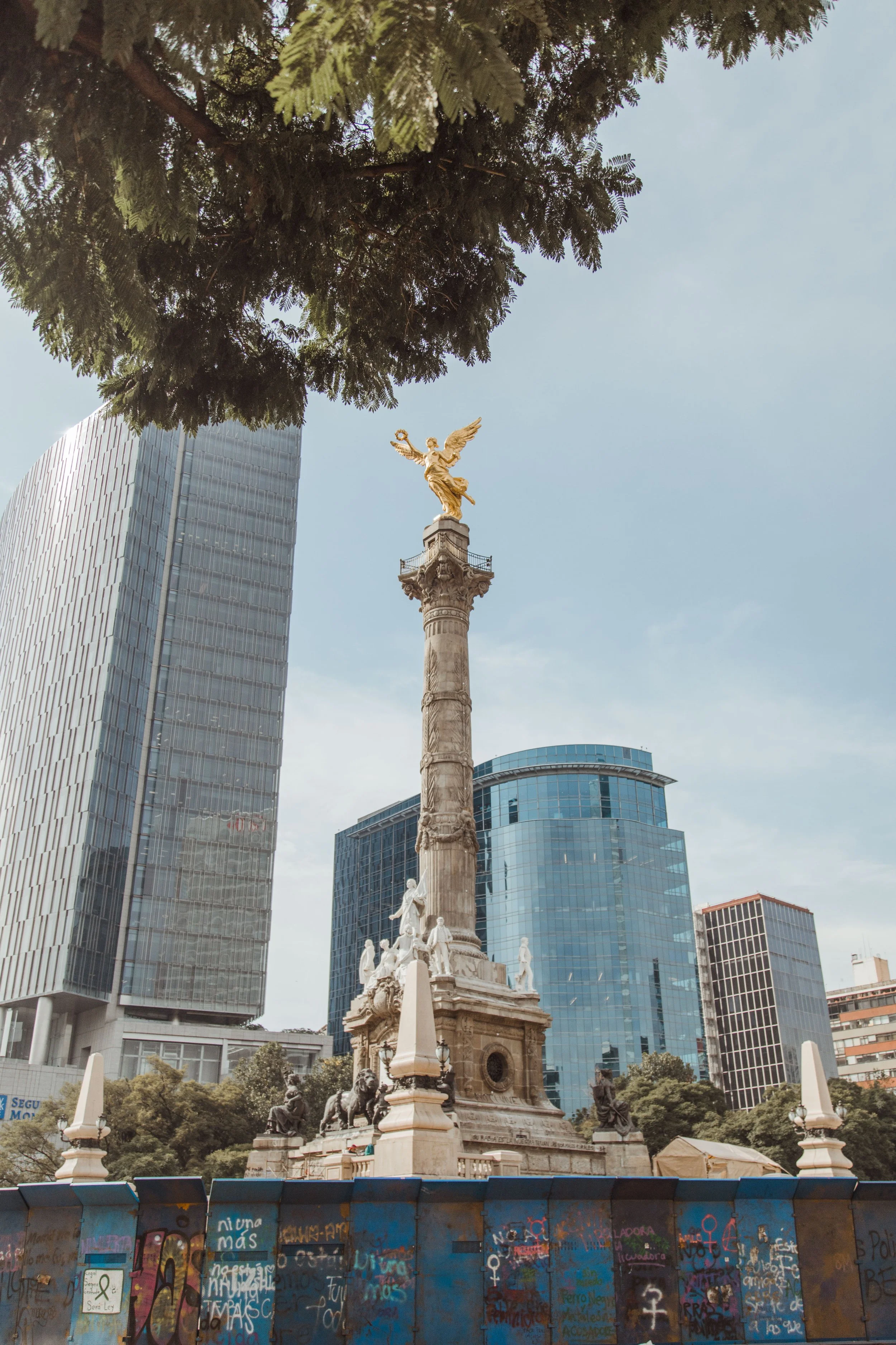 A monument with a tall column topped by a golden statue of Nike, the Greek goddess of victory, in an urban park with modern buildings in the background and graffiti-covered barriers in the foreground.
