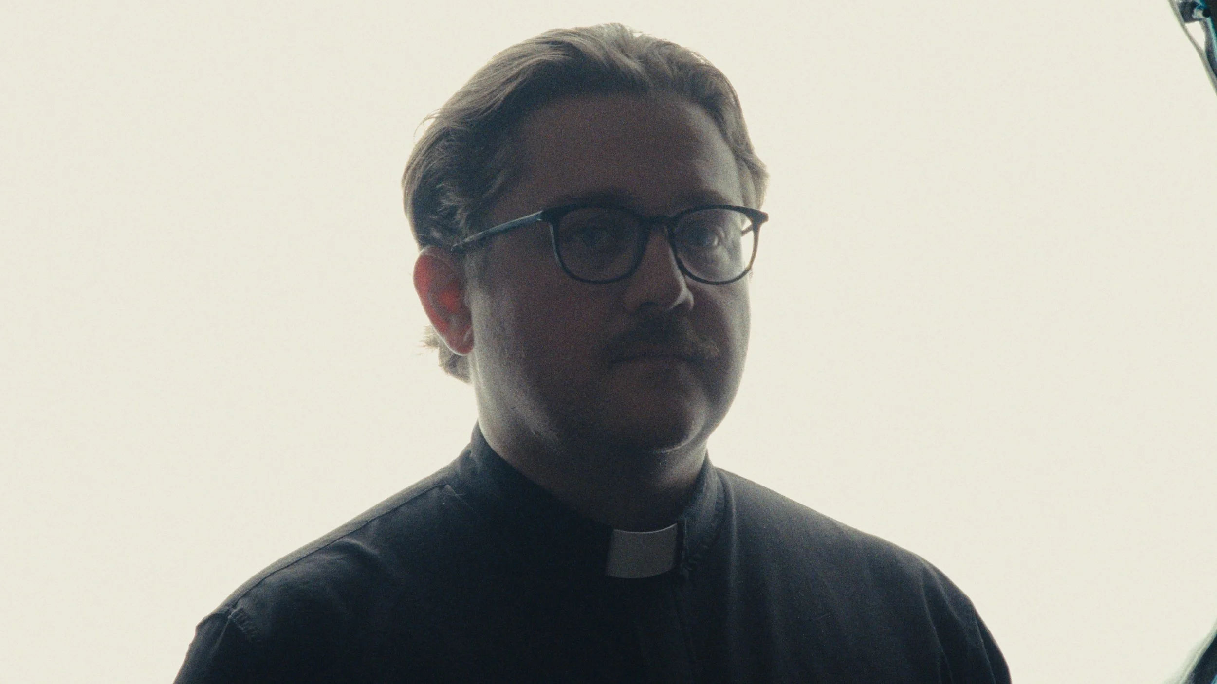 A person dressed as a priest wearing glasses, with light-colored hair, photographed against a plain white background.