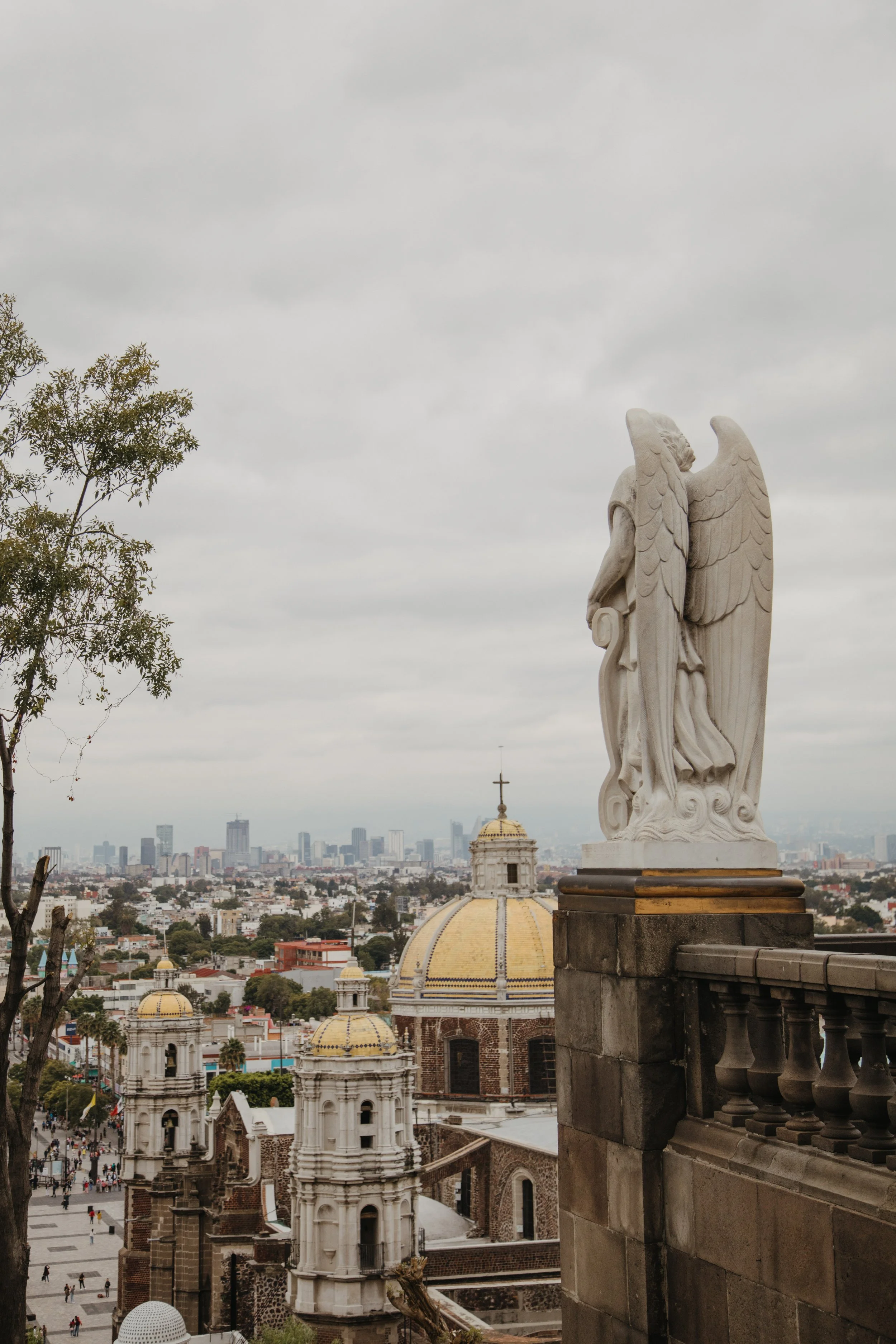 Statue of an angel with wings overlooking a cityscape, including historic buildings with yellow domes and modern skyscrapers in the distance, under a cloudy sky.