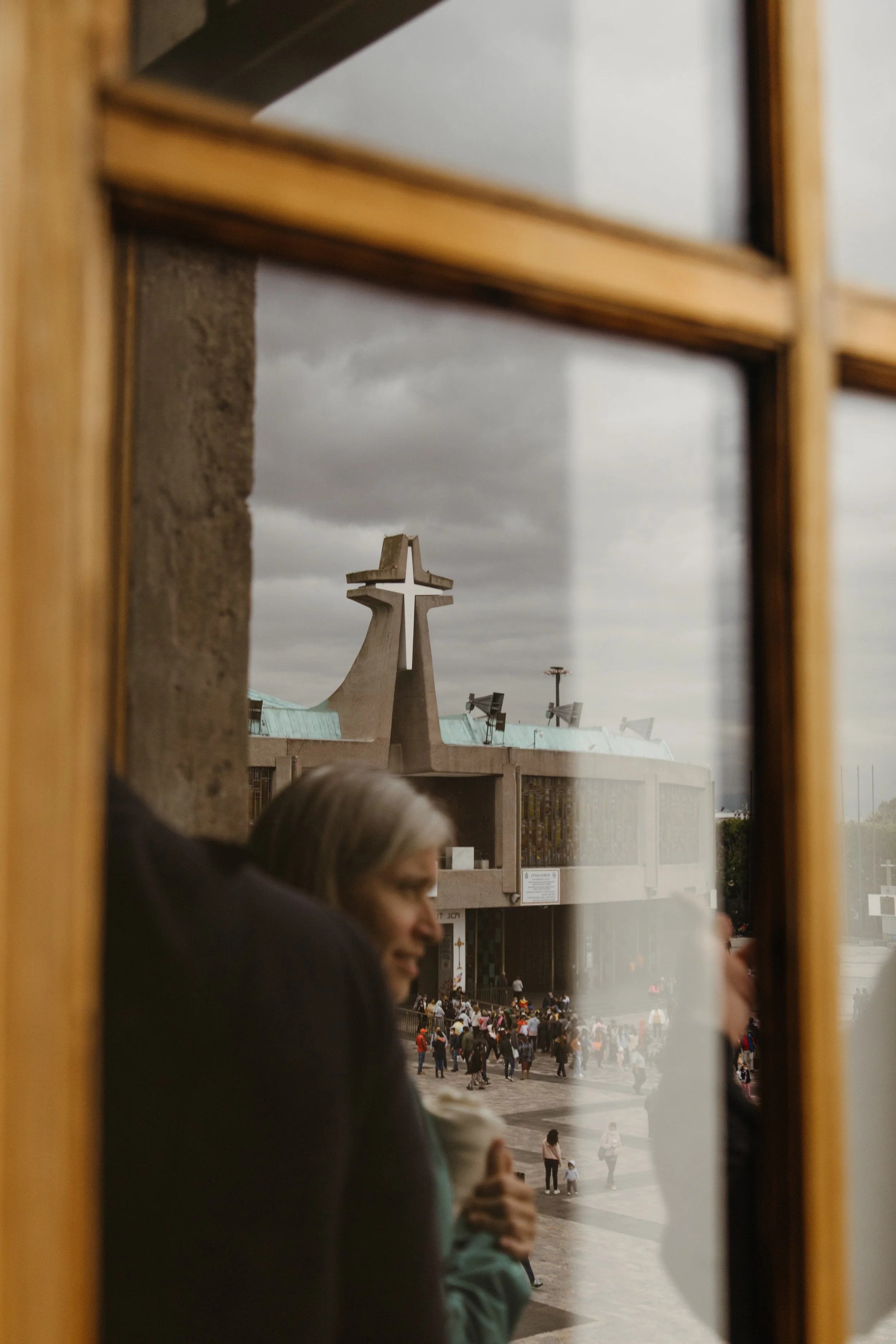 Reflection of a woman holding an ice cream cone seen through a window, with a church or religious building with a cross on its roof in the background, and a crowd of people in an outdoor plaza under cloudy skies.