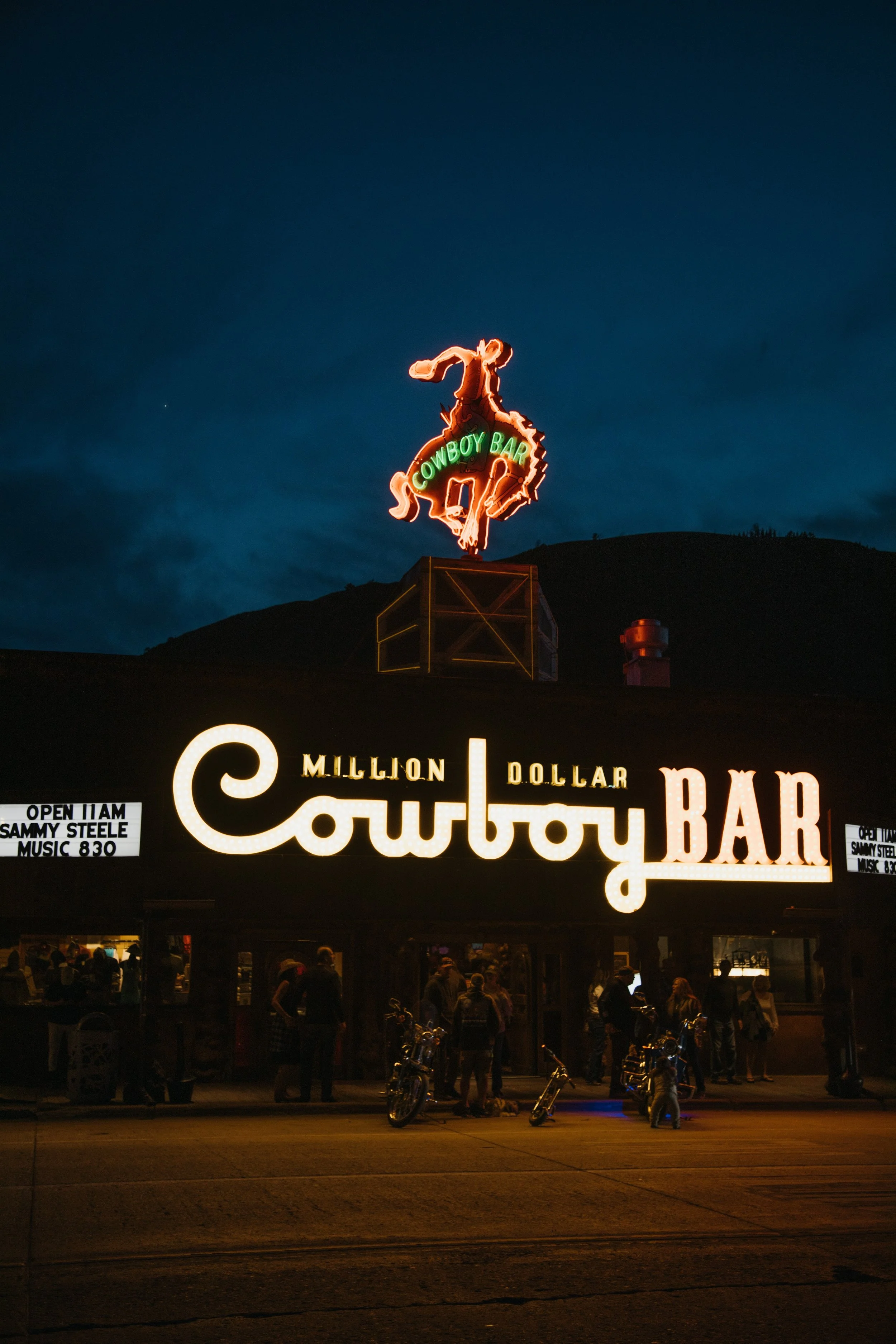 Night view of the Cowboy Bar exterior with illuminated sign and neon cowboy riding horse above, with some people and motorcycles in front.