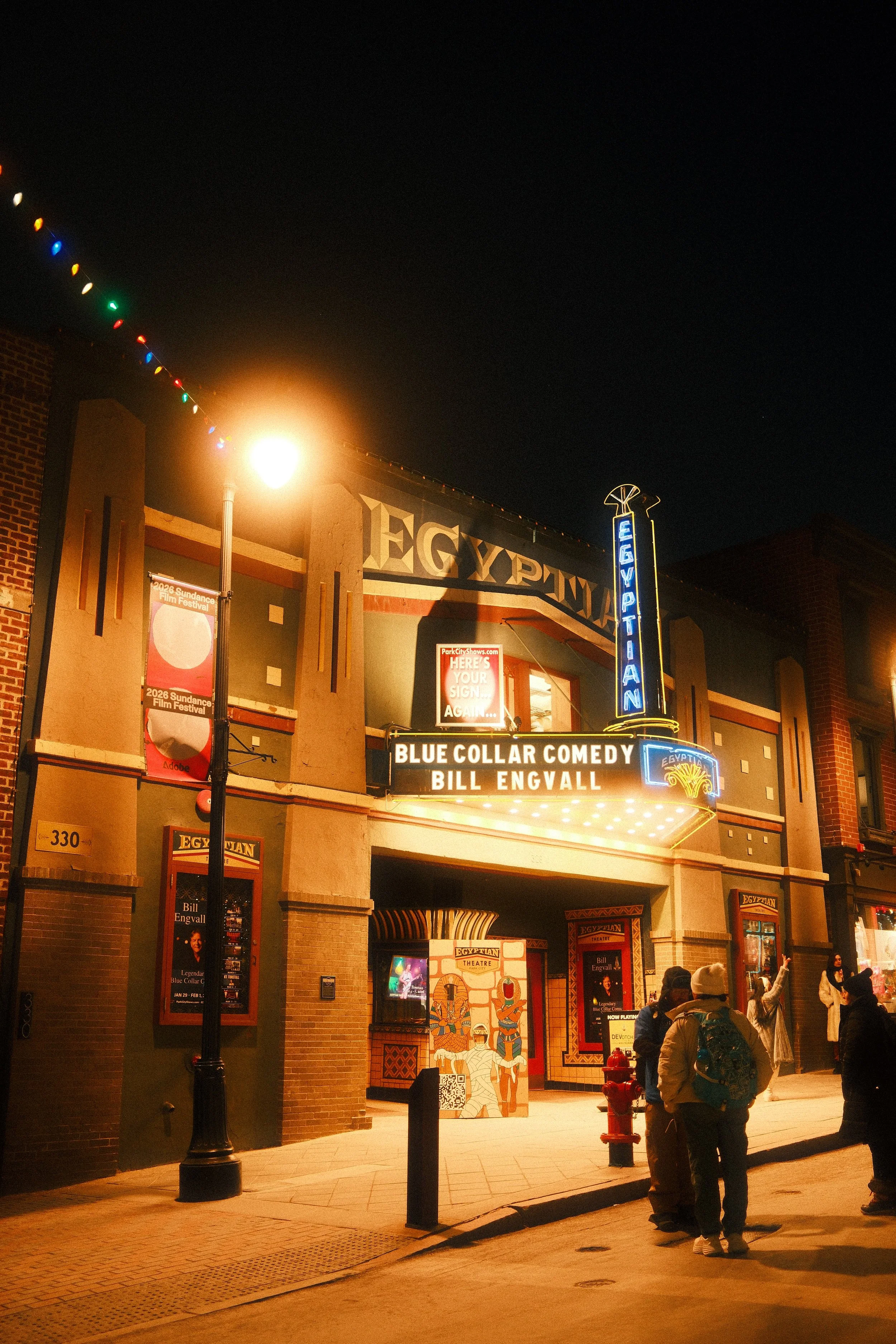 People gathered outside the Egyptian Theatre at night, with neon and marquee signs advertising Bill Engvall's comedy show, and decorative Egyptian-themed artwork near the entrance.