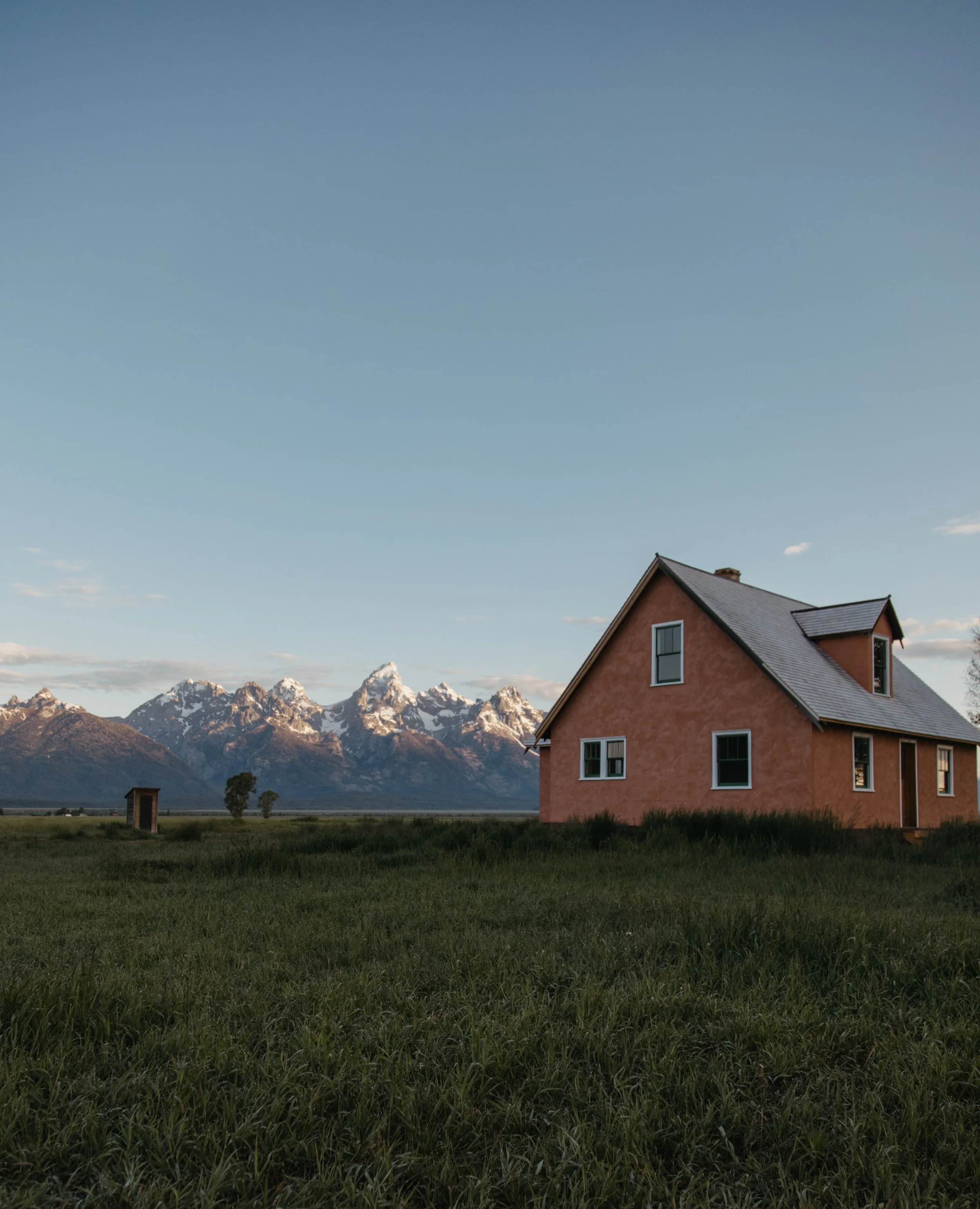 A house with a pink stucco exterior and multiple windows in an open grassy field, mountains with snow-capped peaks in the background, under a clear blue sky.