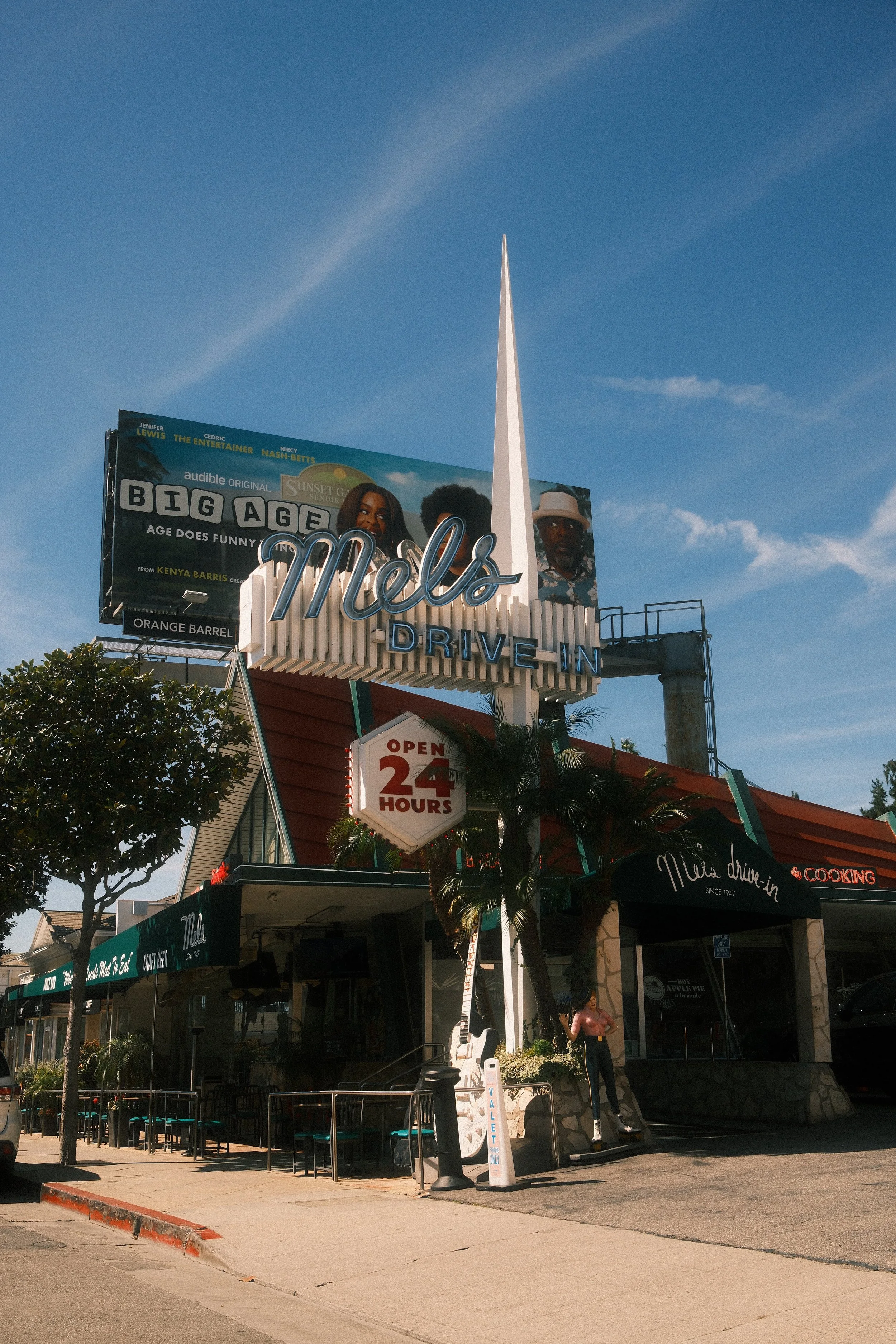 The exterior of Mel's Drive-In fast food restaurant with a large neon sign, palm trees, a person skateboarding, and a billboard in the background under a clear blue sky.