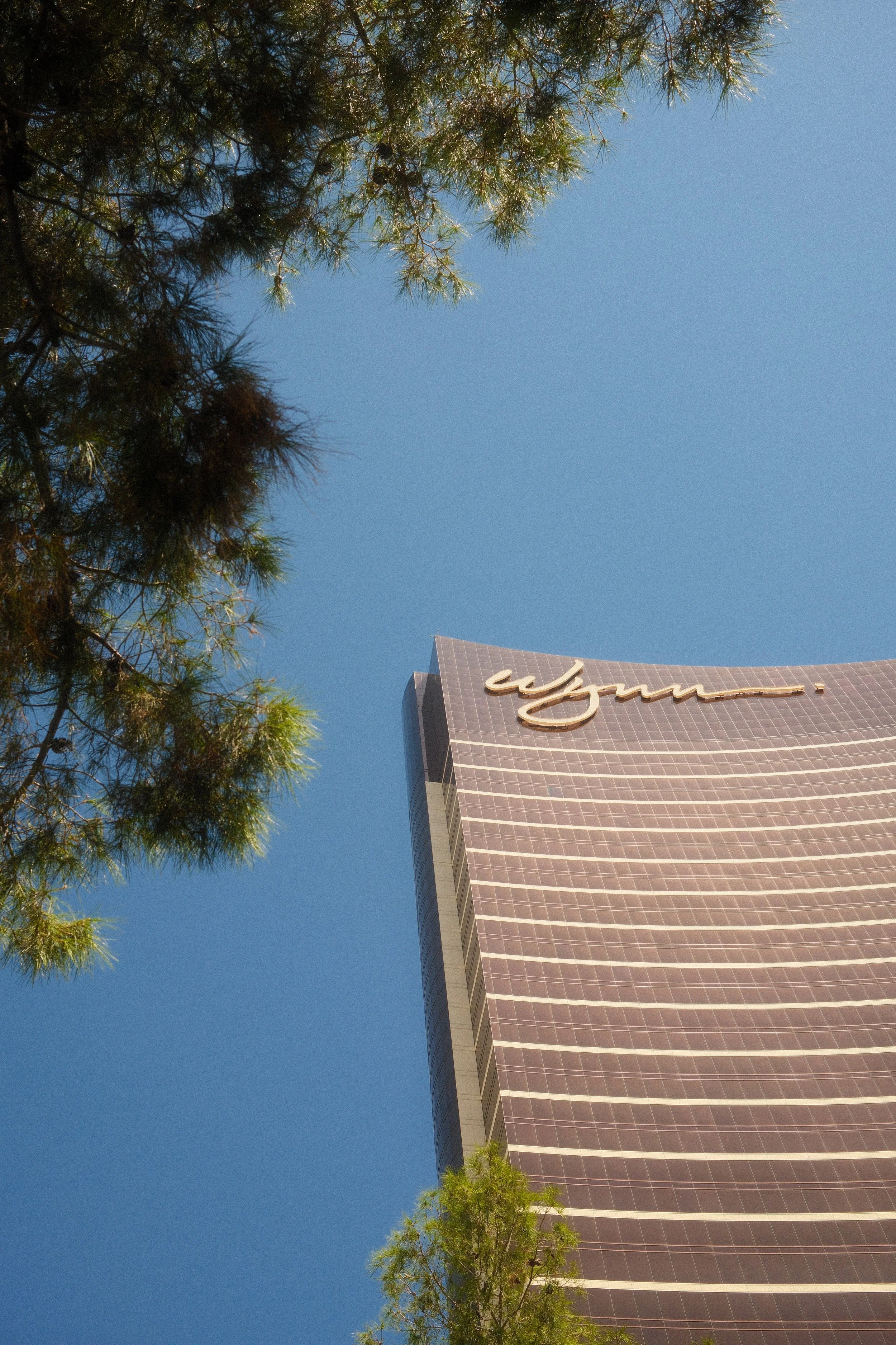 Looking up at a Wynn hotel skyscraper with a blue sky and green trees in the foreground.