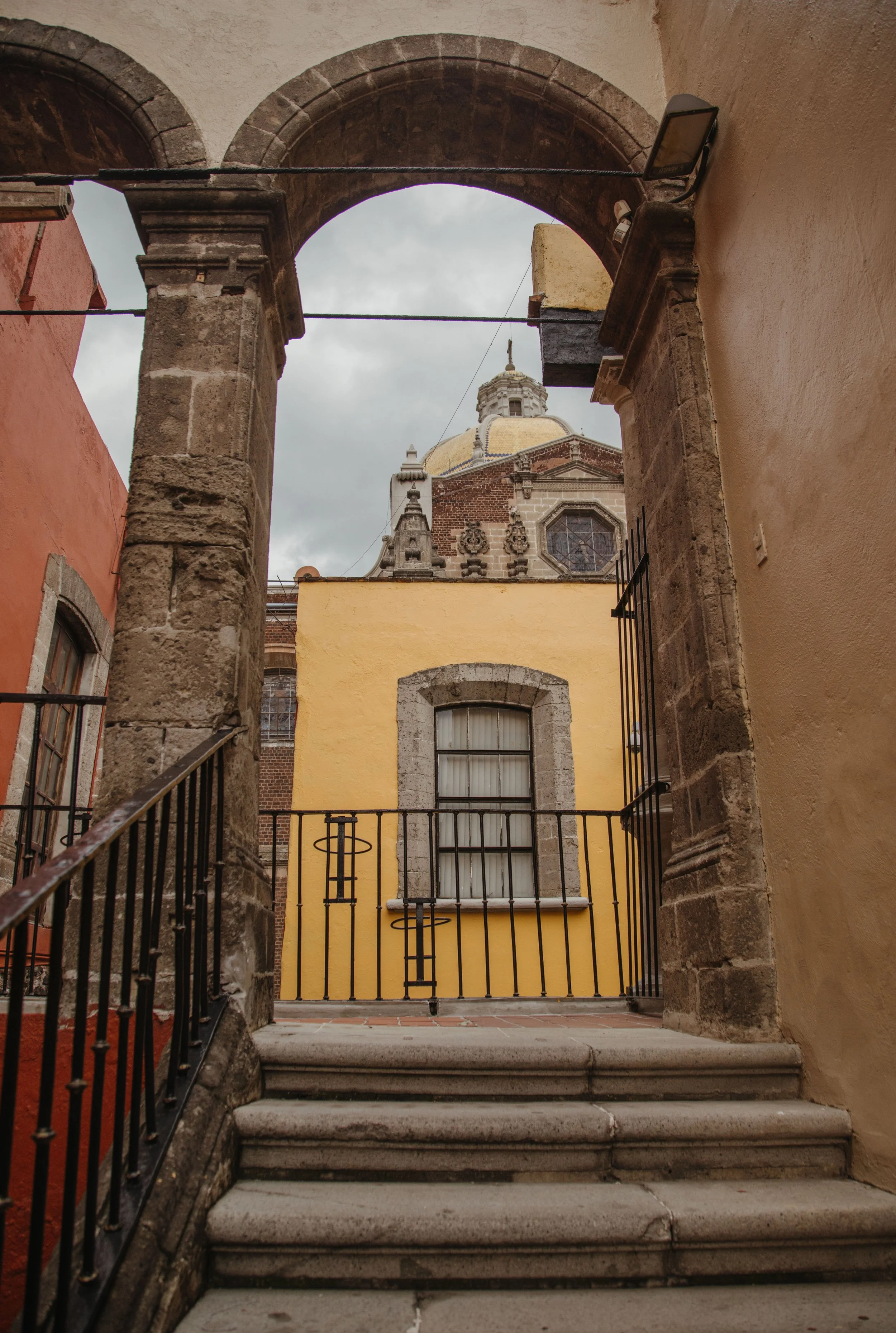 View through a stone archway showing a yellow building with a barred window and a church with a dome and ornate architectural details in the background.