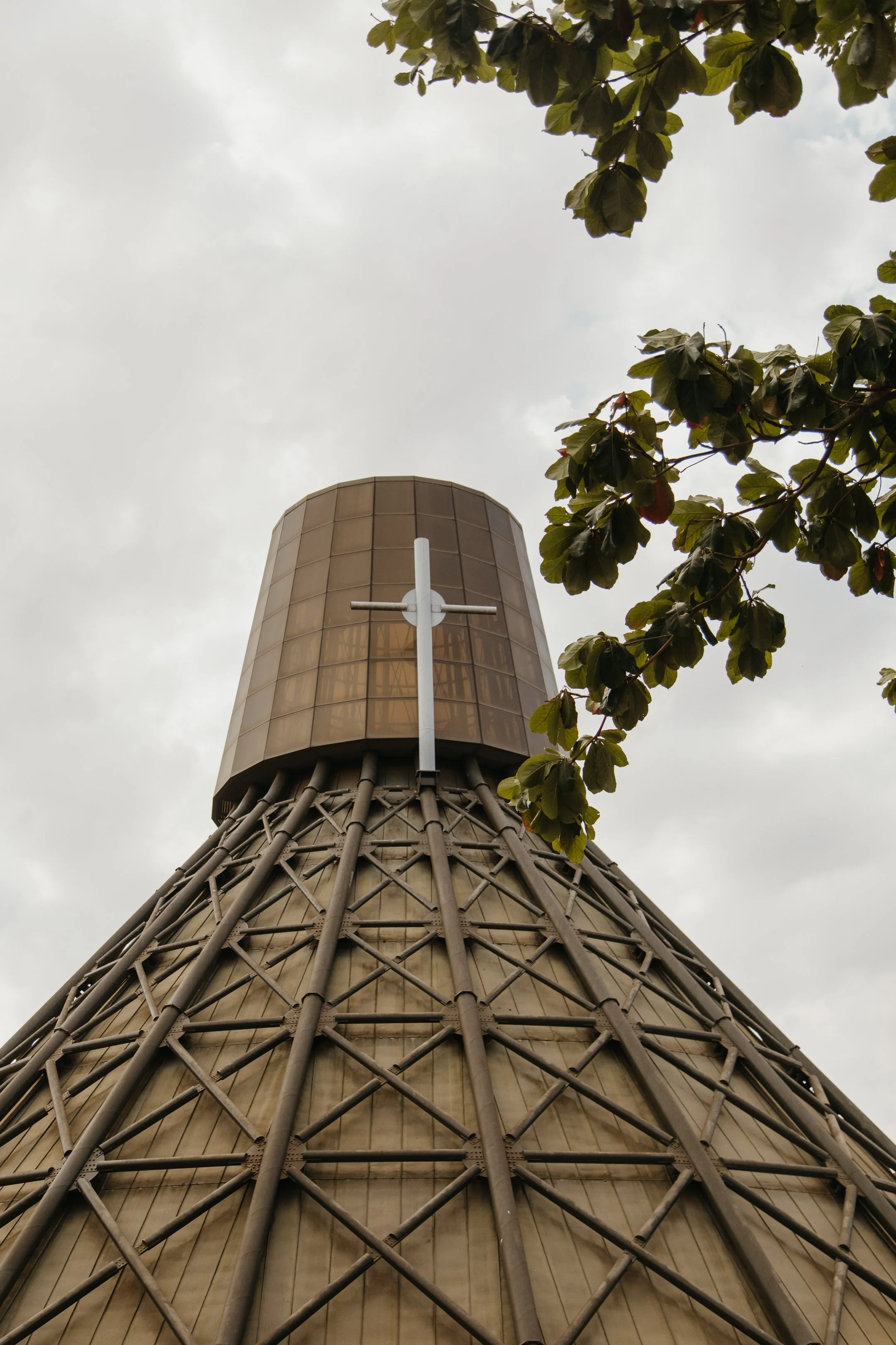 Low-angle view of a large church steeple with a cross, partly obscured by tree leaves against a cloudy sky.