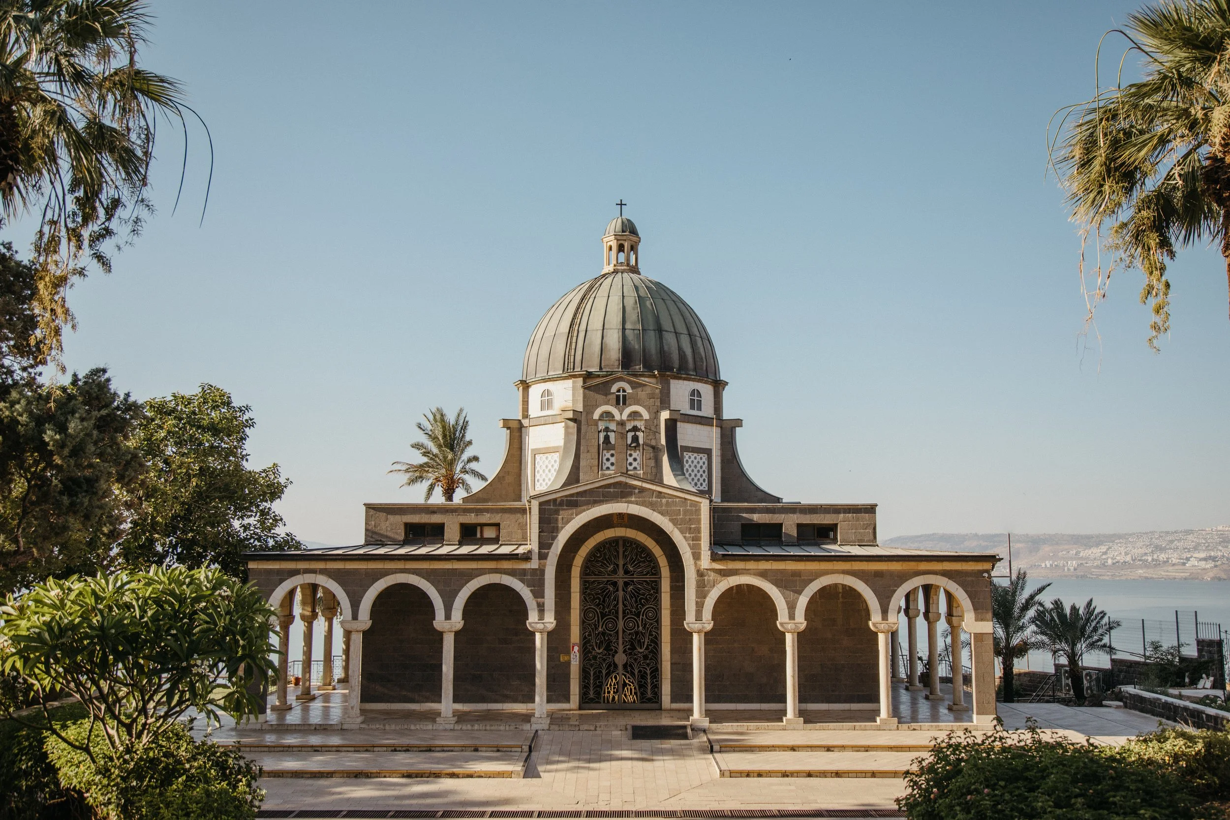 A church with a large central dome, arched entrance, and surrounding columns, set against a clear blue sky with trees and water in the background.