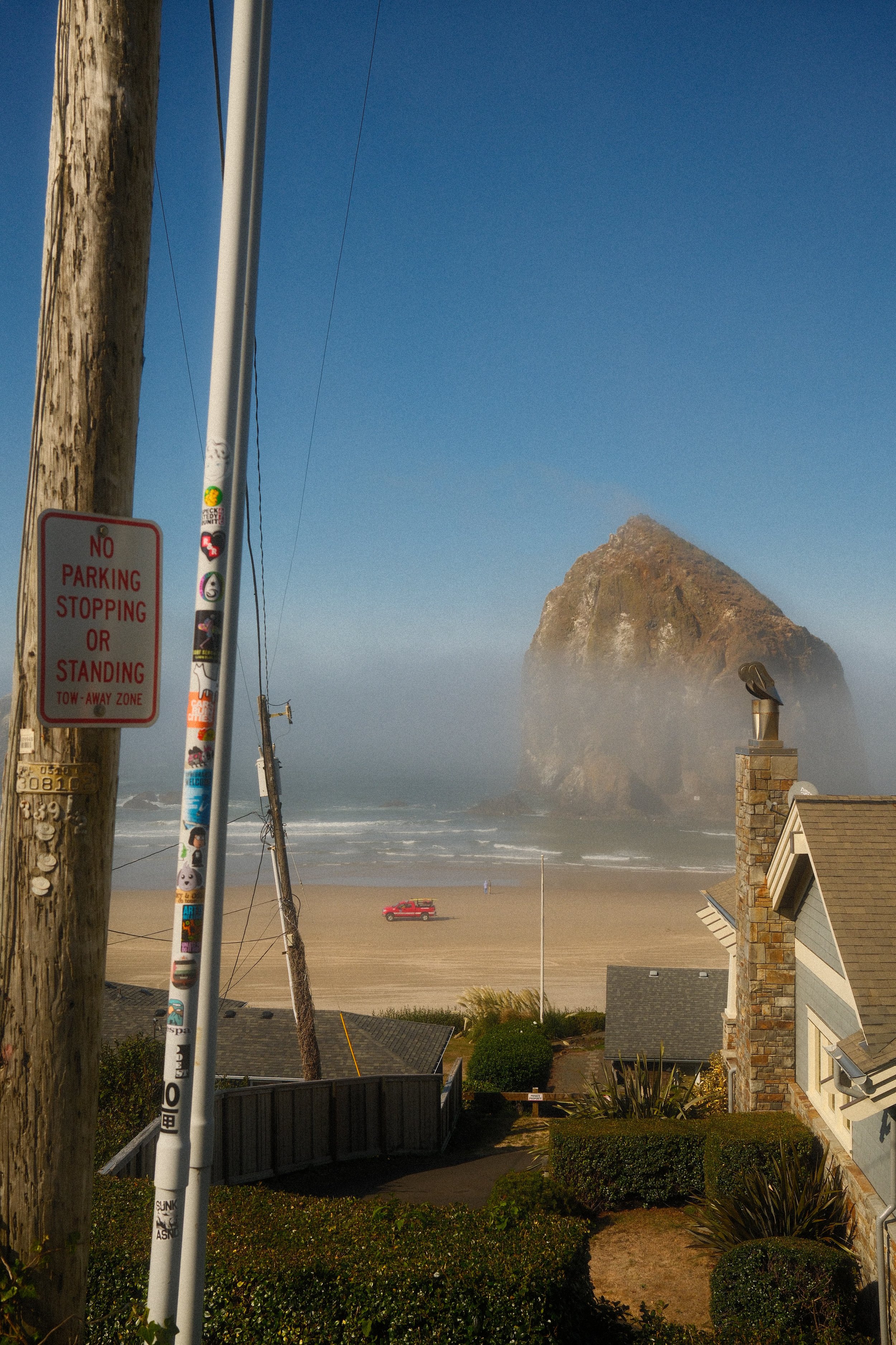 A beach scene featuring large sea stacks, a sandy shore, and the ocean with waves. Foreground includes residential rooftops, a utility pole, and a no parking or standing sign.