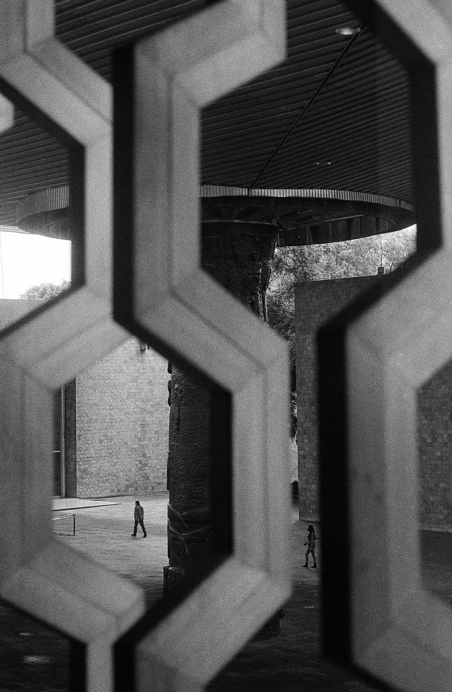 View of a person walking outdoors through a decorative geometric cutout in a metal fence or gate, in black and white.