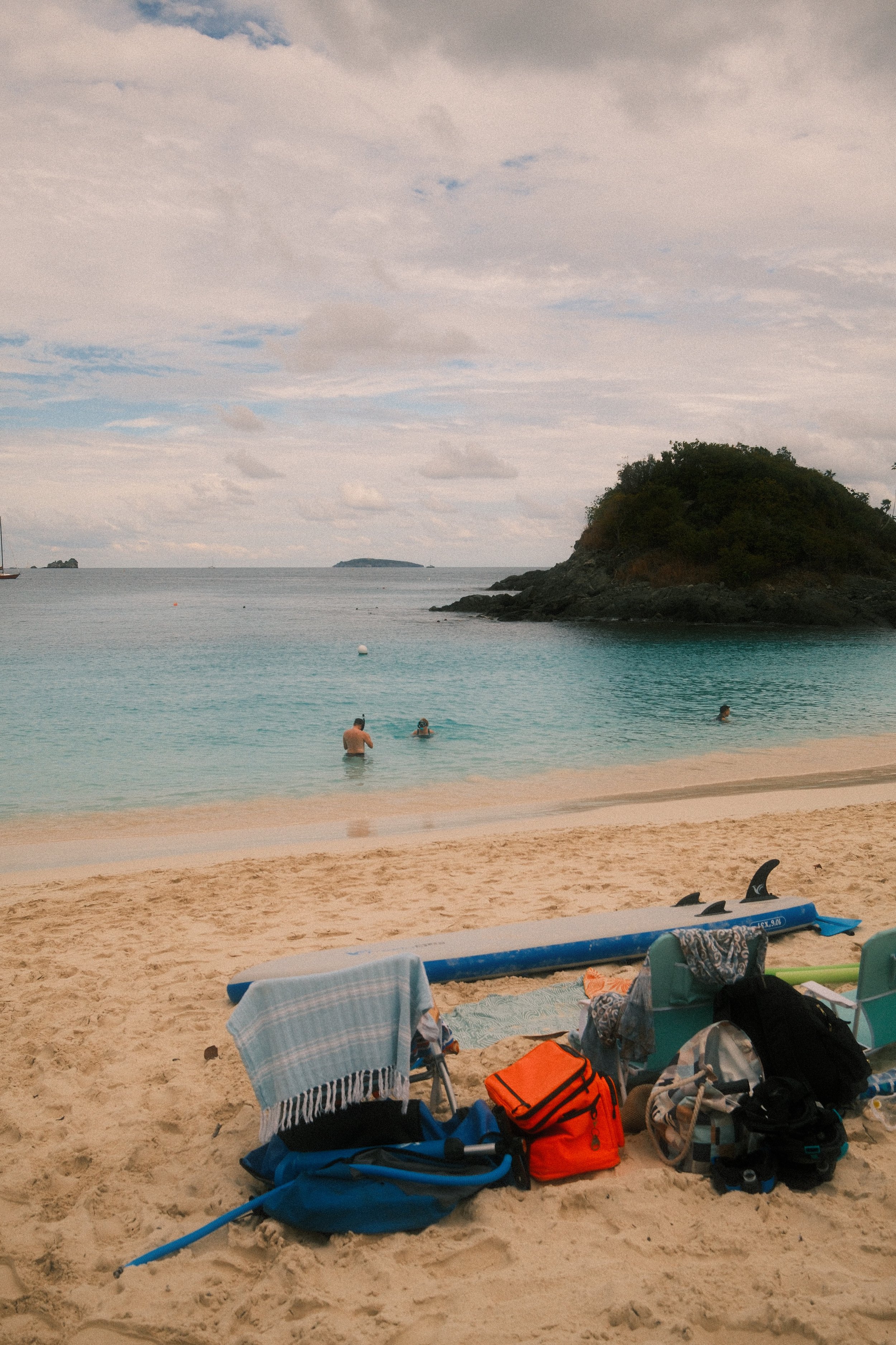 A beach scene with a sandy shore, surfboards, and beach gear in the foreground. People are swimming in the calm, turquoise water, with an island and a boat in the background. Overcast sky with clouds.