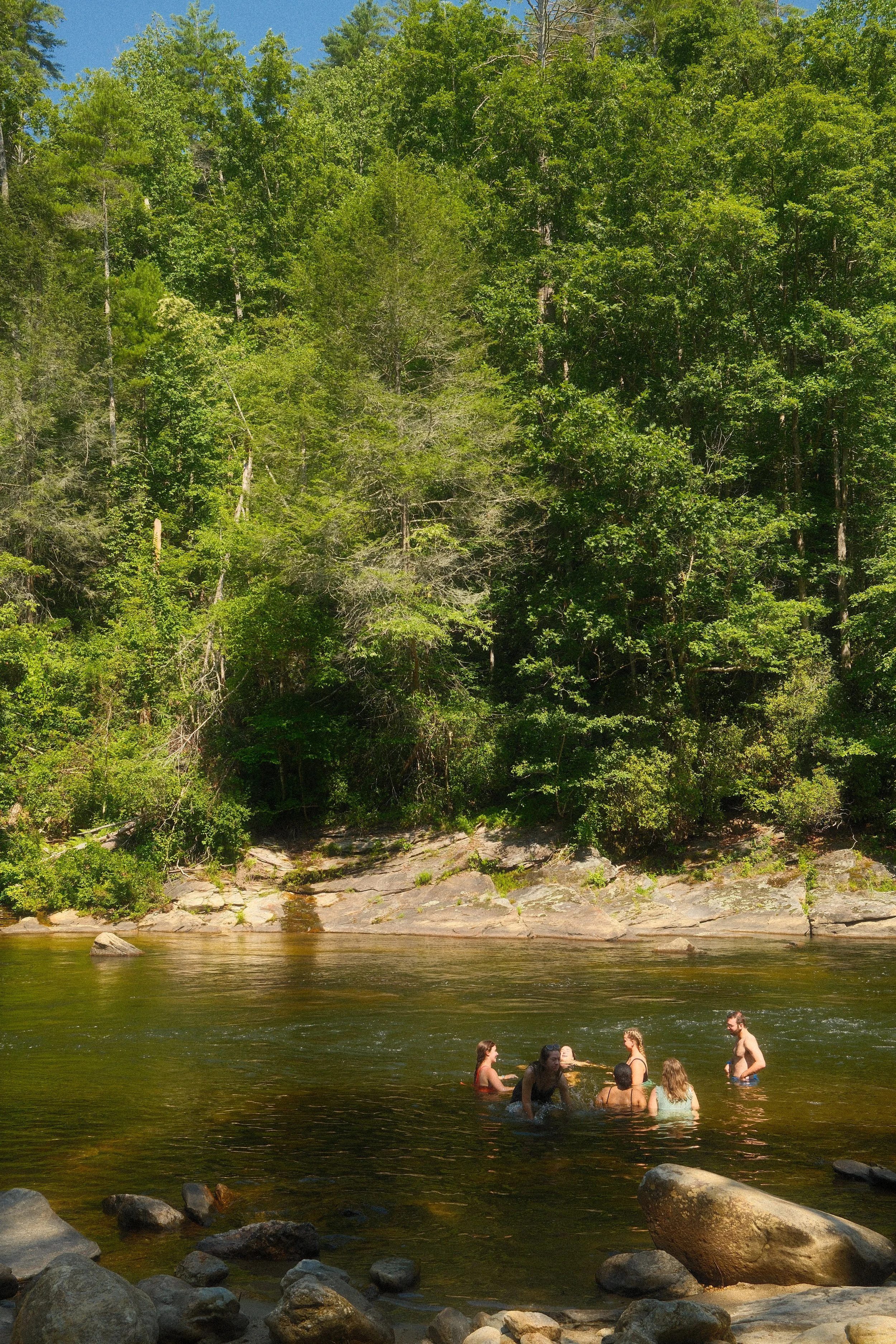 People swimming and relaxing in a natural river surrounded by lush green trees and large rocks under a clear blue sky.