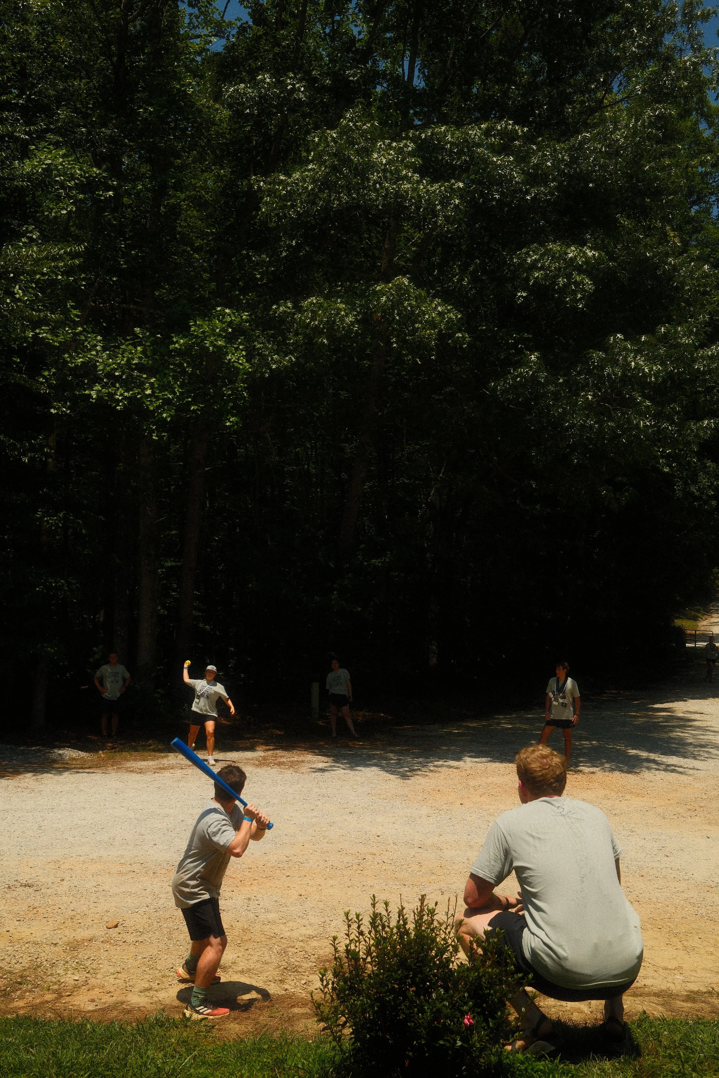 Children playing baseball outdoors on a dirt field with trees in the background, one child swinging a blue bat, others on the field, and an adult sitting on the grass watching.