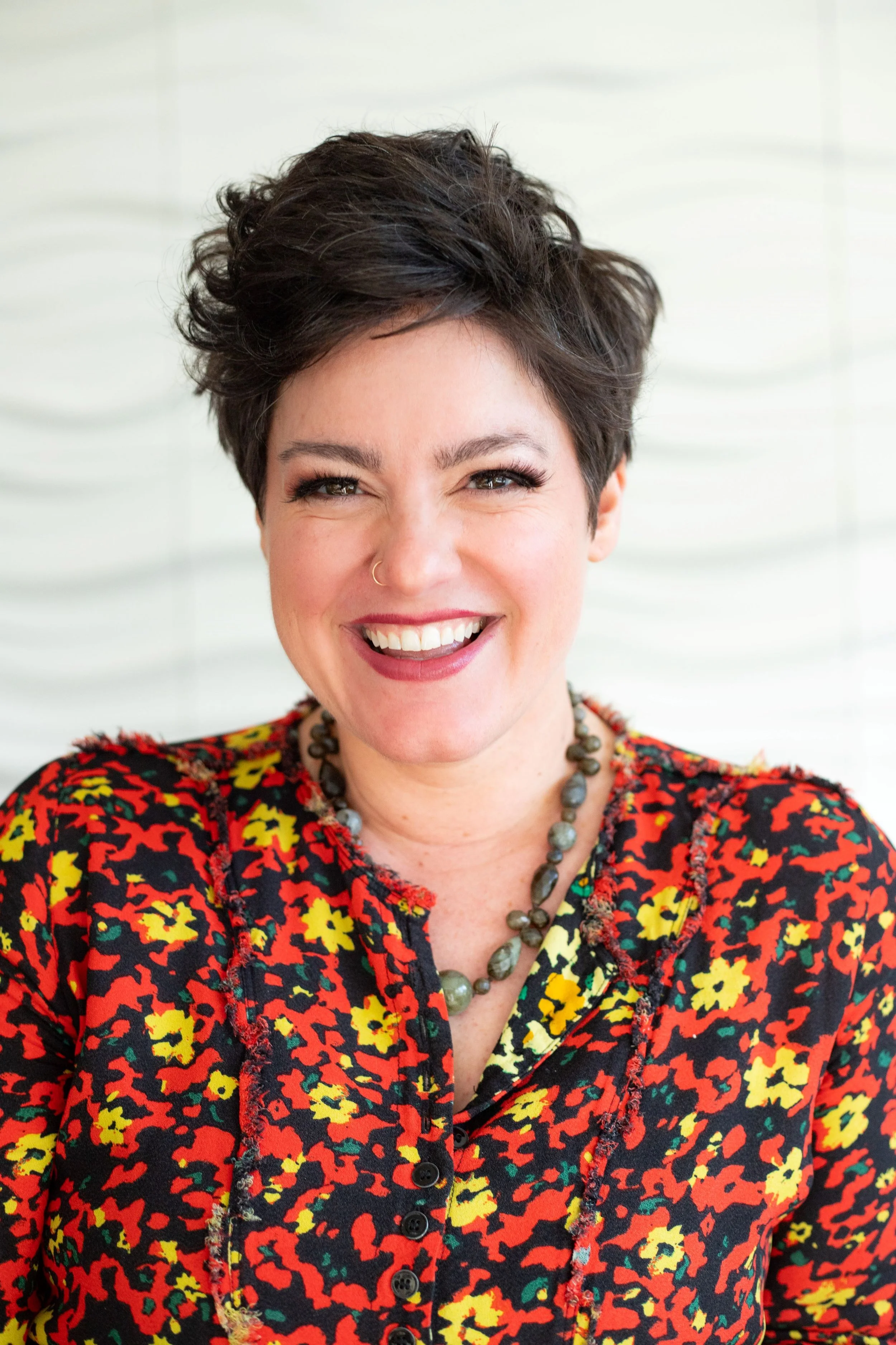 A woman with short, dark, curly hair smiling and wearing a colorful floral blouse and a beaded necklace, standing in front of a white textured background.