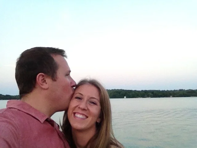 A man kissing a woman on the cheek by a lake, with a sunset sky and trees in the background.