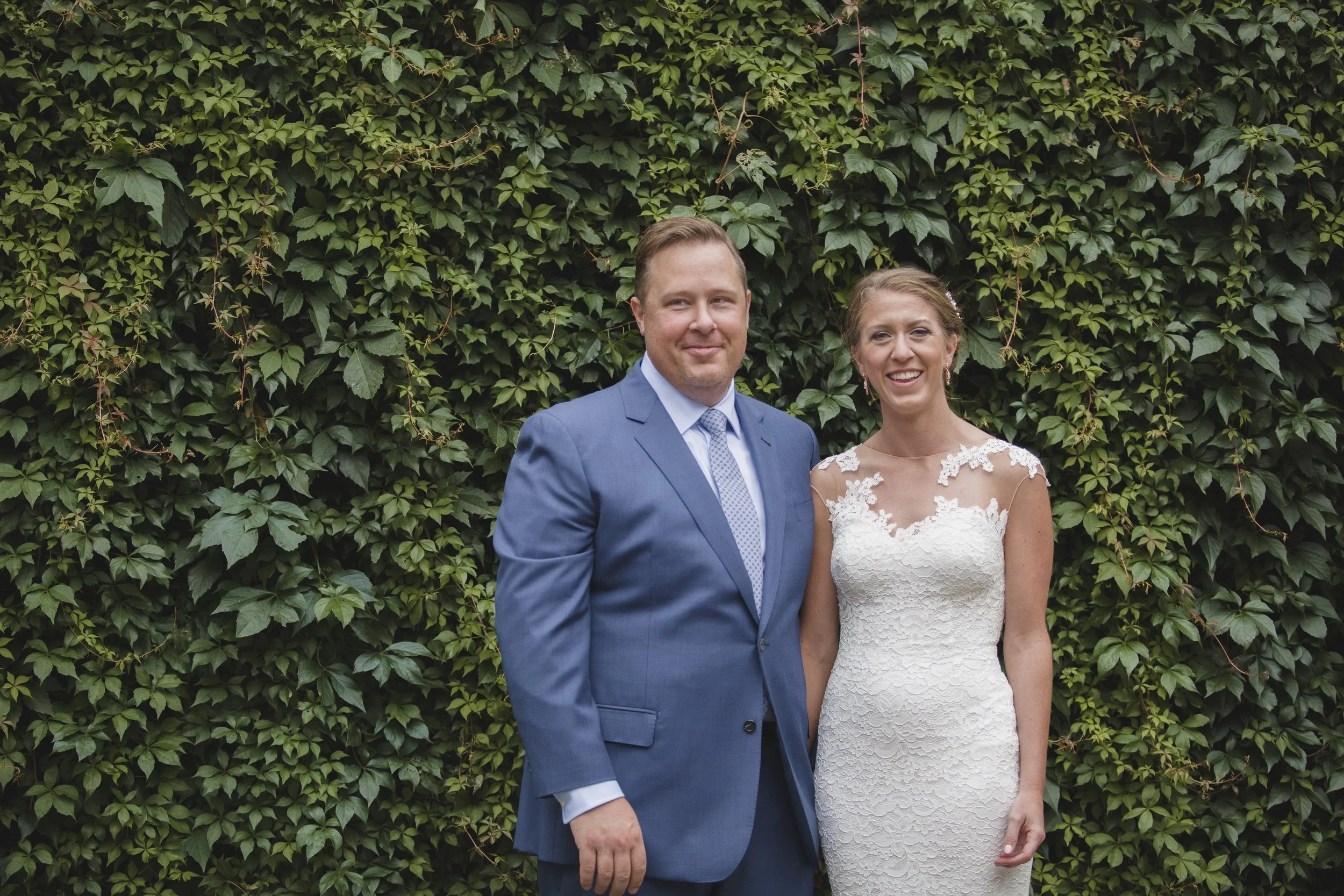 A newlywed couple standing in front of a green ivy-covered wall, the groom in a blue suit and the bride in a white lace wedding dress, both smiling.