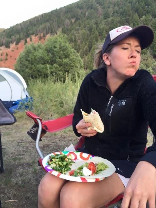 Person sitting outdoors next to a tent and a camping chair, making a facial expression while holding a half-eaten wrap, with a plate of salad and salsa on their lap in a natural setting with trees and hills in the background.