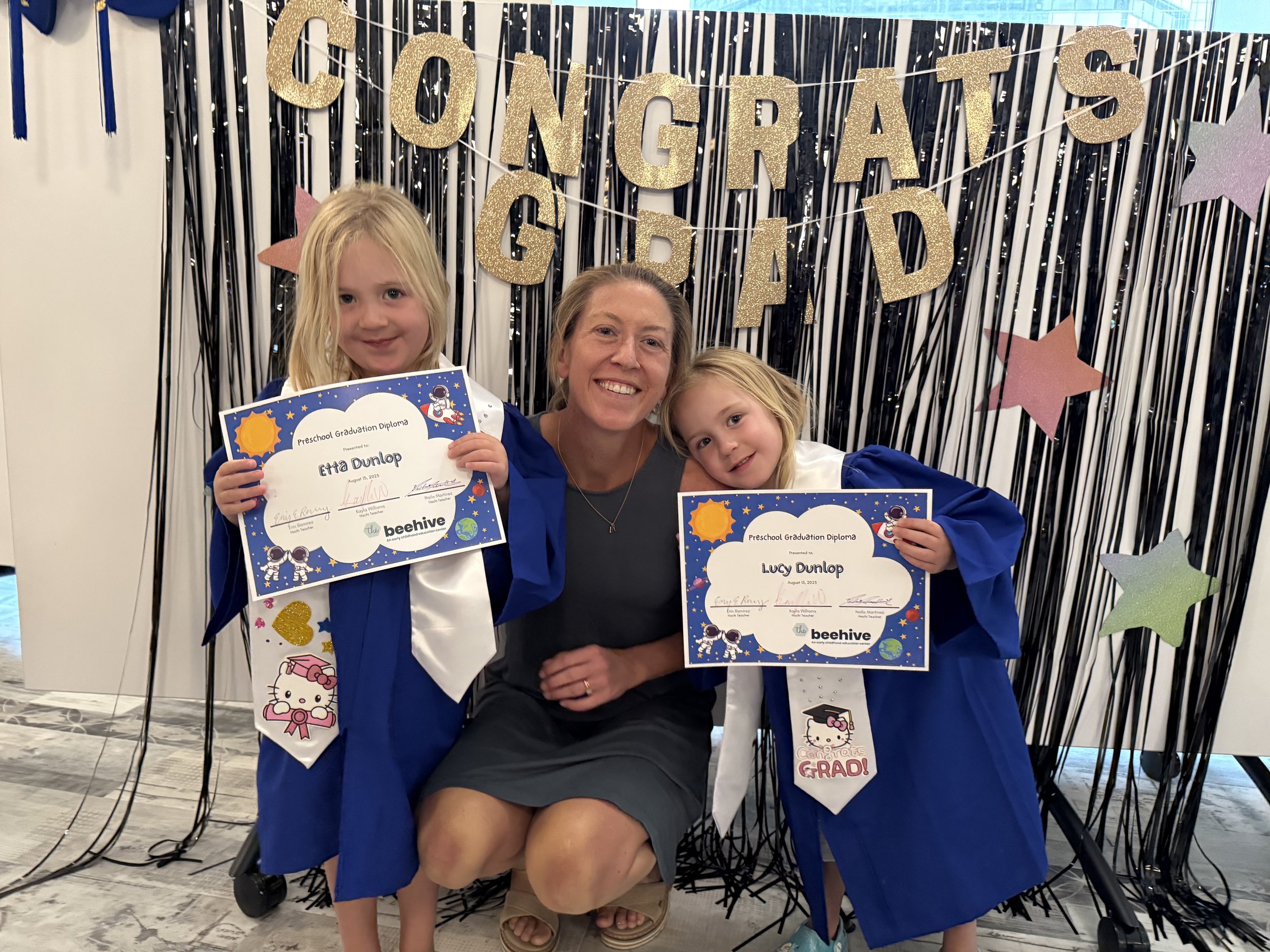 Two young girls in blue graduation gowns holding certificates, with a smiling woman in the middle, at a preschool graduation celebration. Behind them is a decorated backdrop with gold and pastel-colored stars and a banner that says 'CONGRATS GARD'.